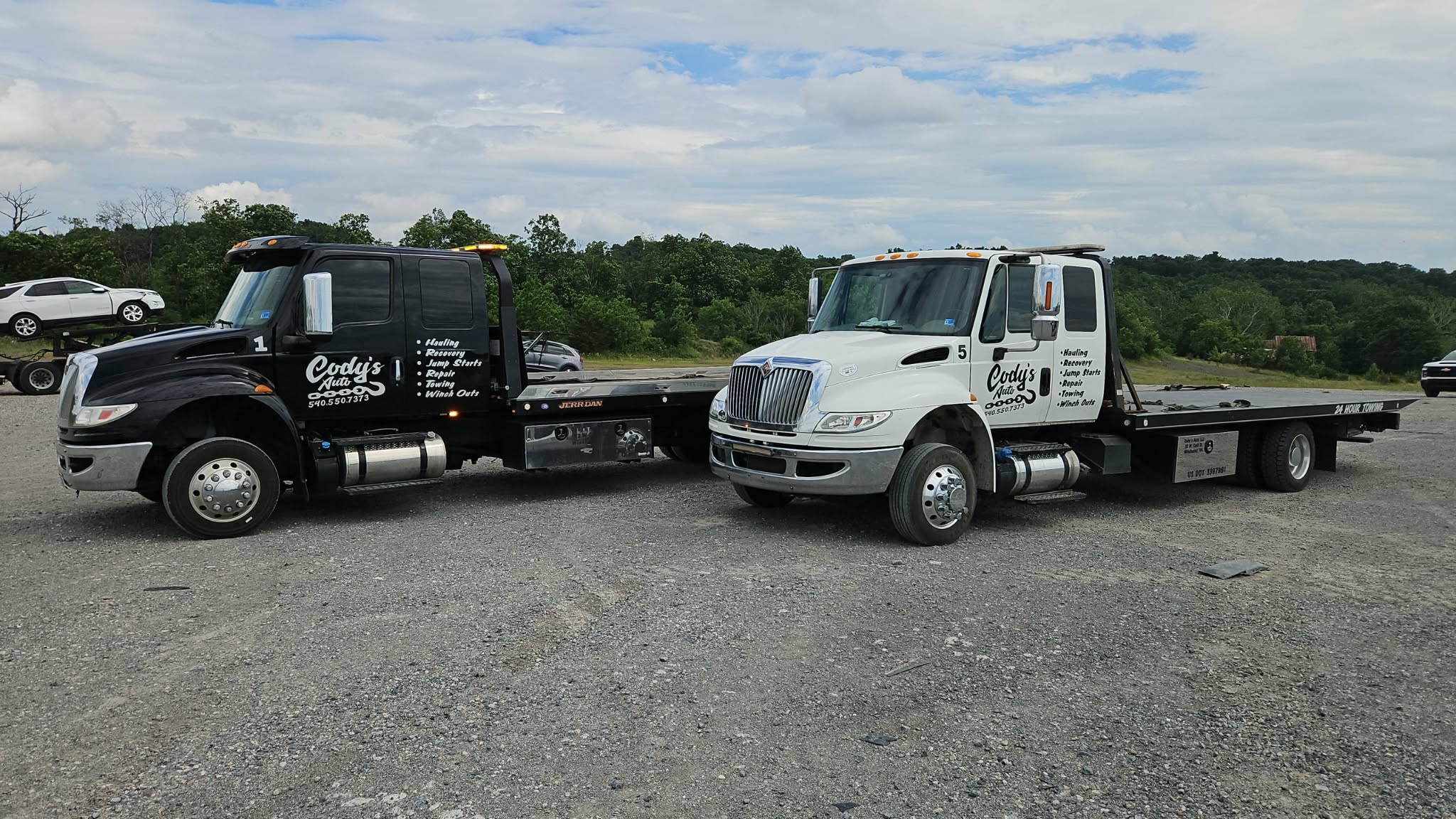 Two flatbed trucks parked side by side on a gravel lot with trees and sky in background.