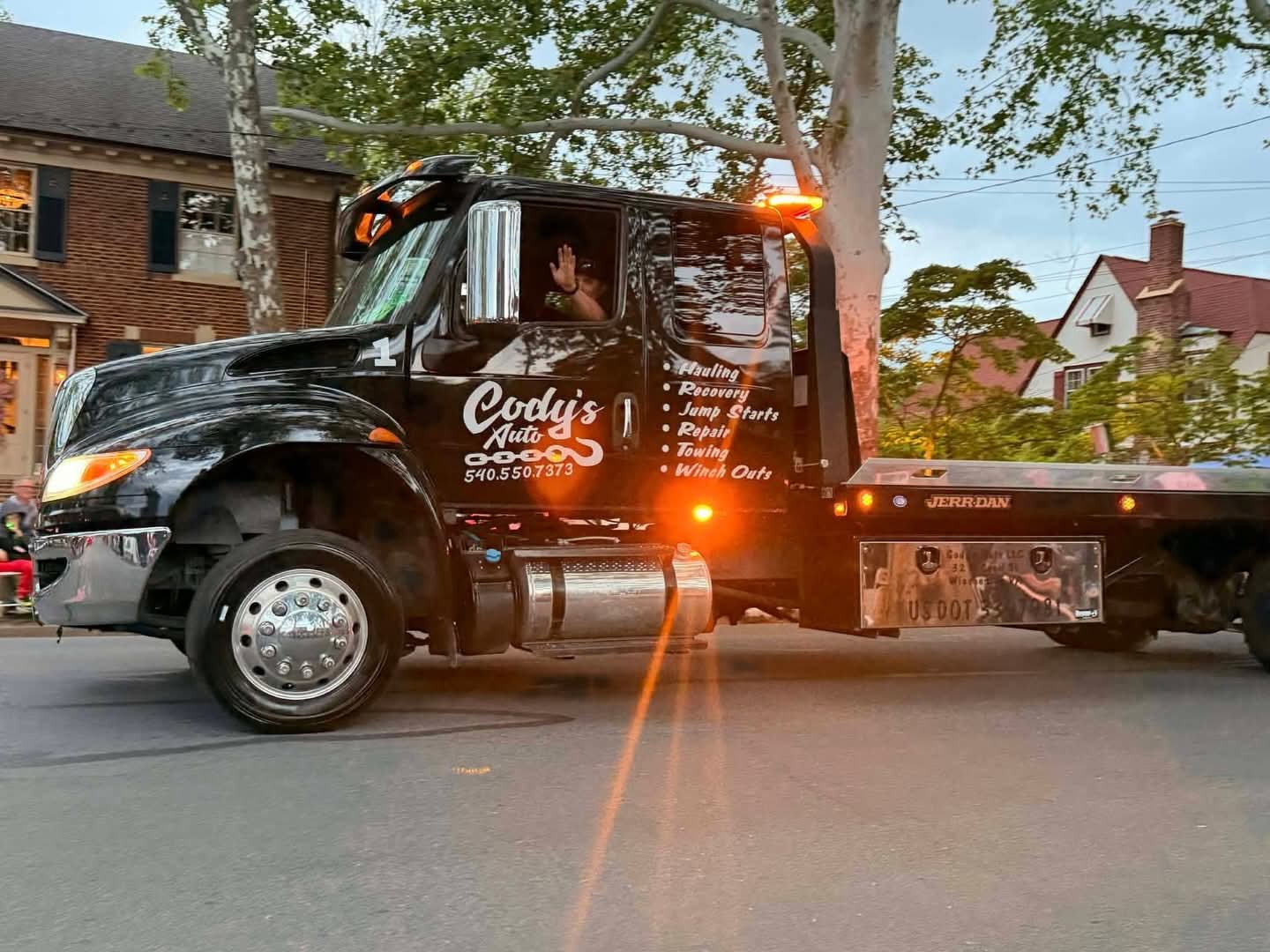 Black tow truck parked on street with driver inside, trees and houses in background.
