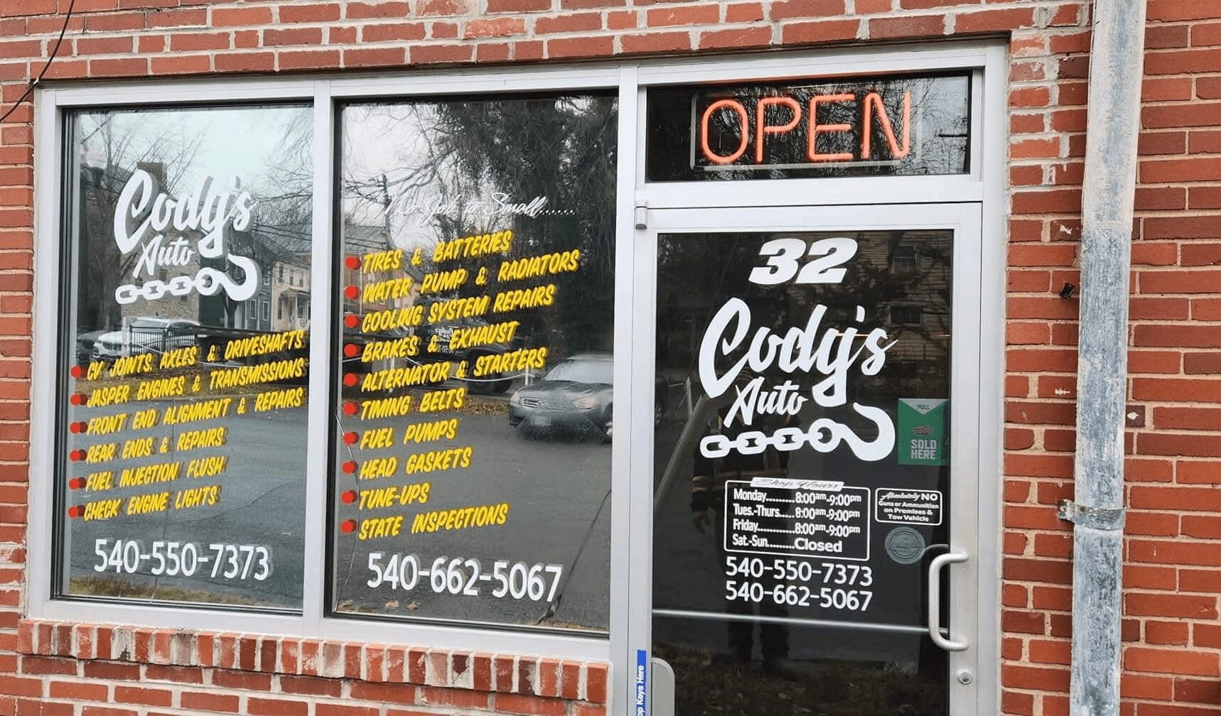 Brick storefront with large windows, neon 'OPEN' sign, and signage for Cody's Auto, including phone numbers.