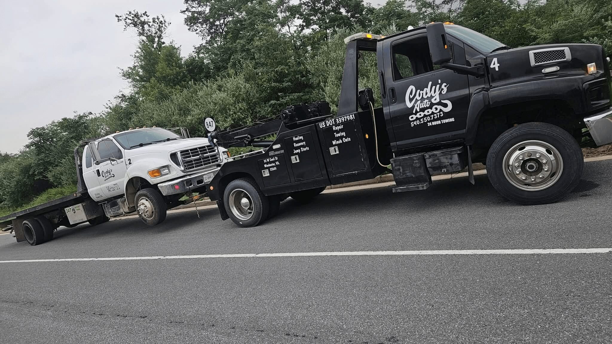 Tow truck with a white car on its flatbed, parked on the side of a road with trees in the background.