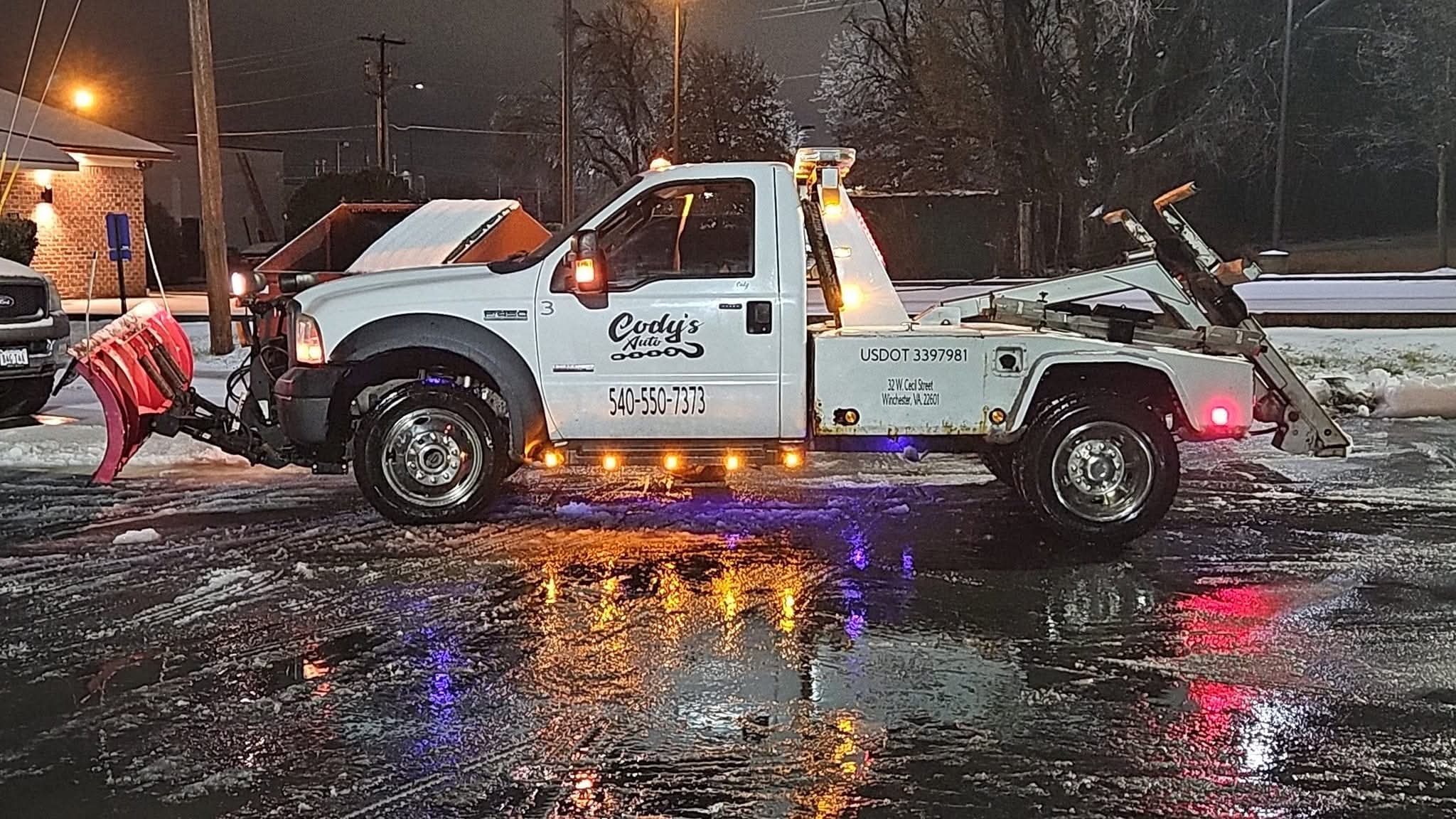 Damaged tow truck on wet road with debris and reflective water, nighttime scene with trees and buildings in background.