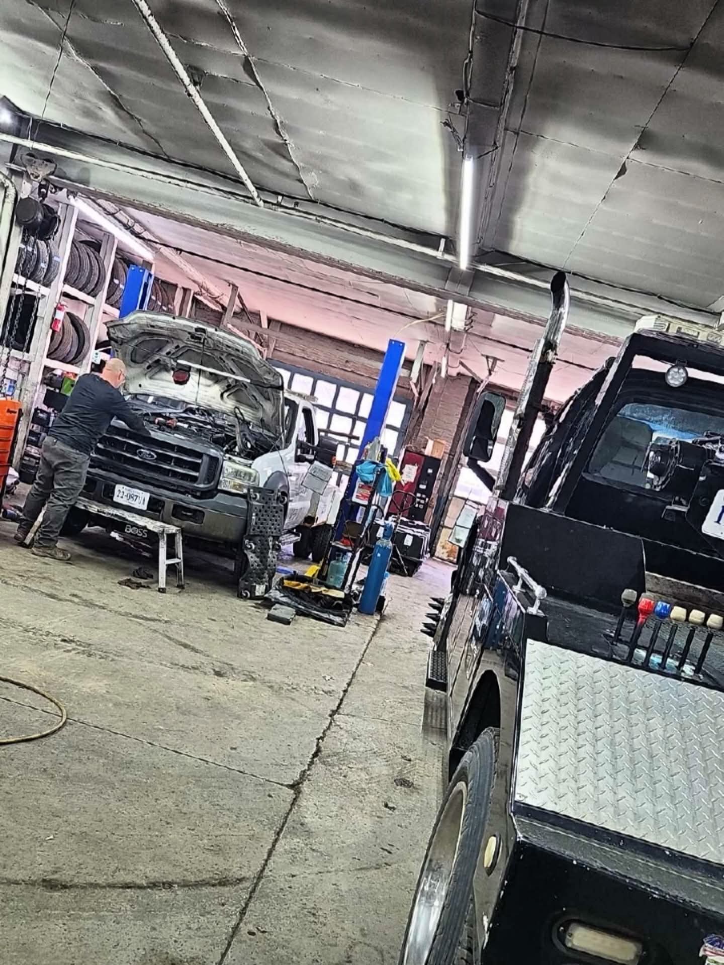 Automotive repair shop with vehicles, tools, and a person working on a car, ceiling pipes, and equipment visible.