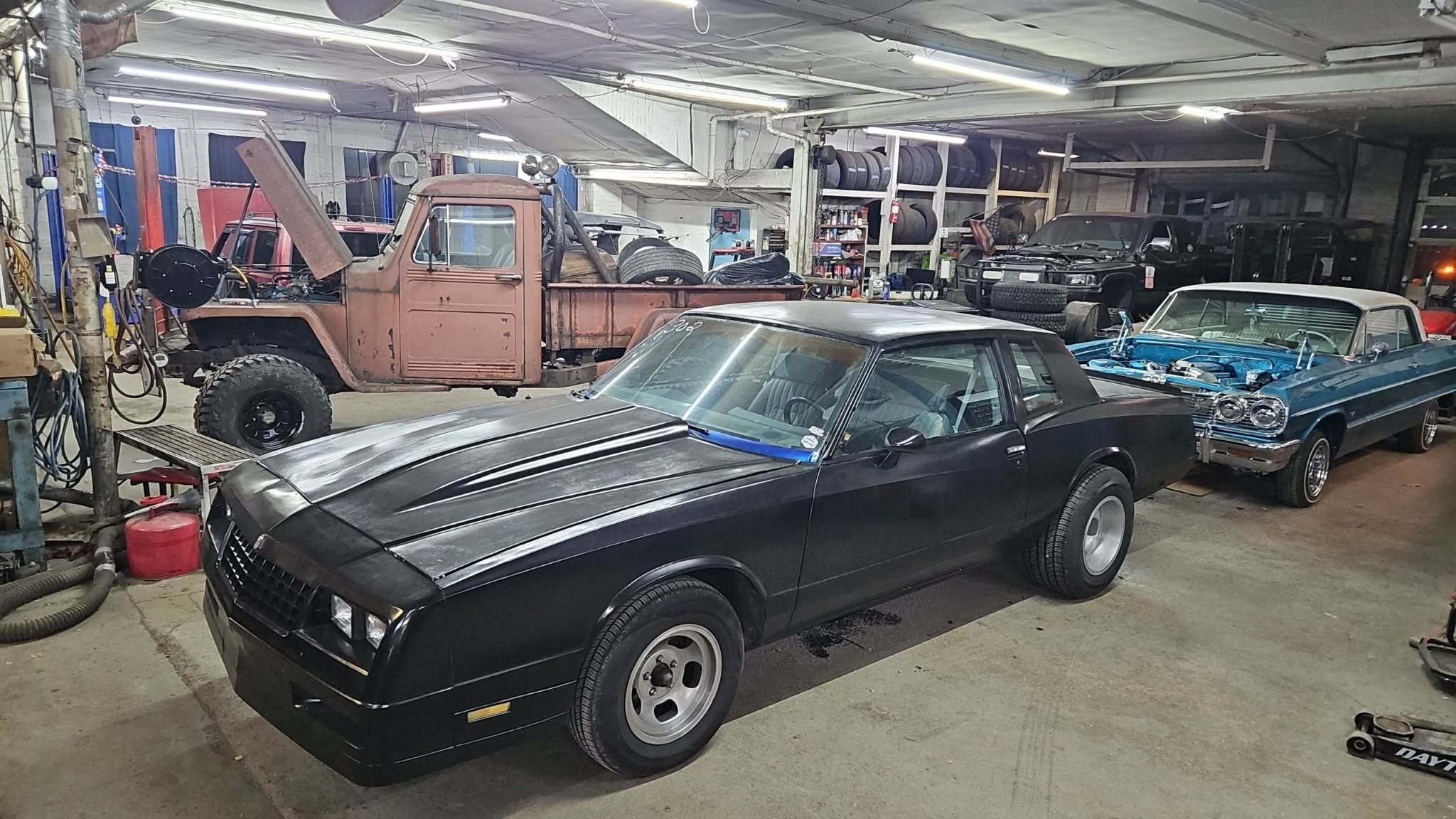 Black vintage car parked indoors with other classic vehicles and equipment in the background.