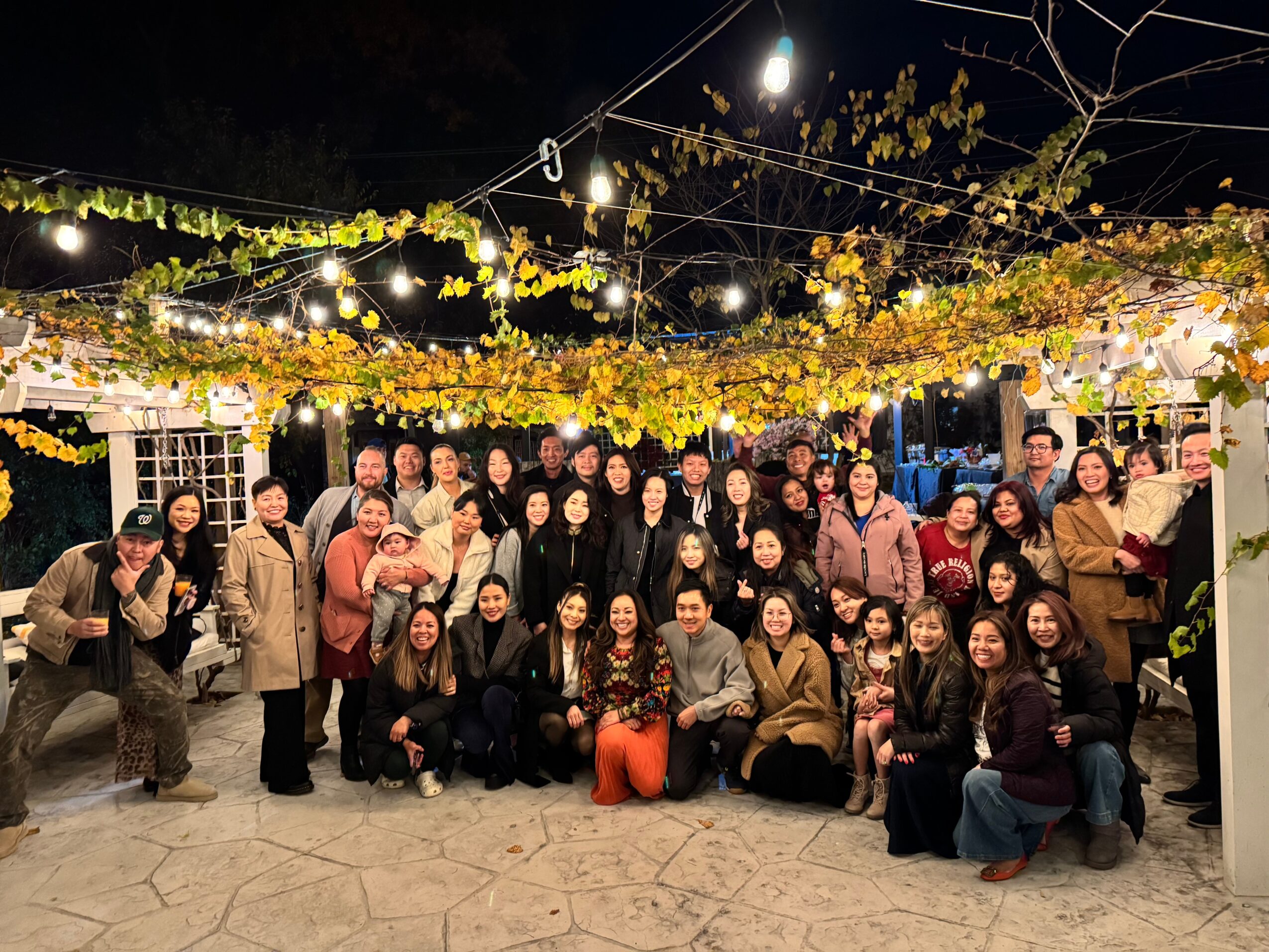Group of people gathered outdoors at night under string lights and yellow foliage, posing for a photo.