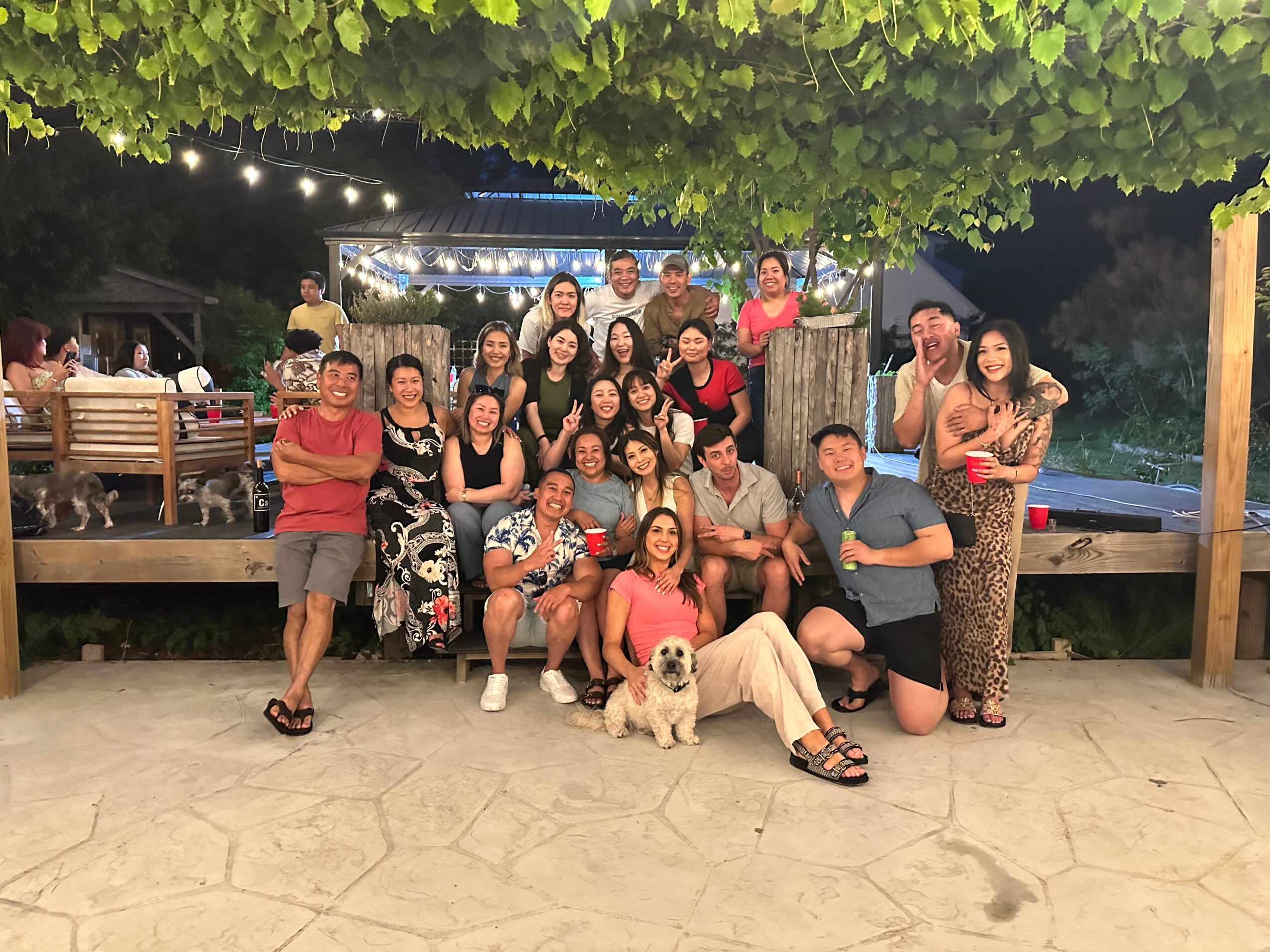 Group of people gathered outdoors at night, standing and sitting, with a dog in front, under a leafy canopy.