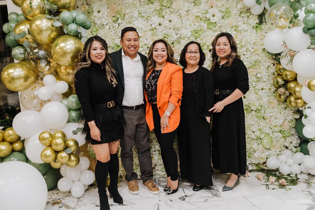 Five people standing in front of a floral and balloon backdrop, smiling, dressed in black, orange, and gray, celebrating an event.