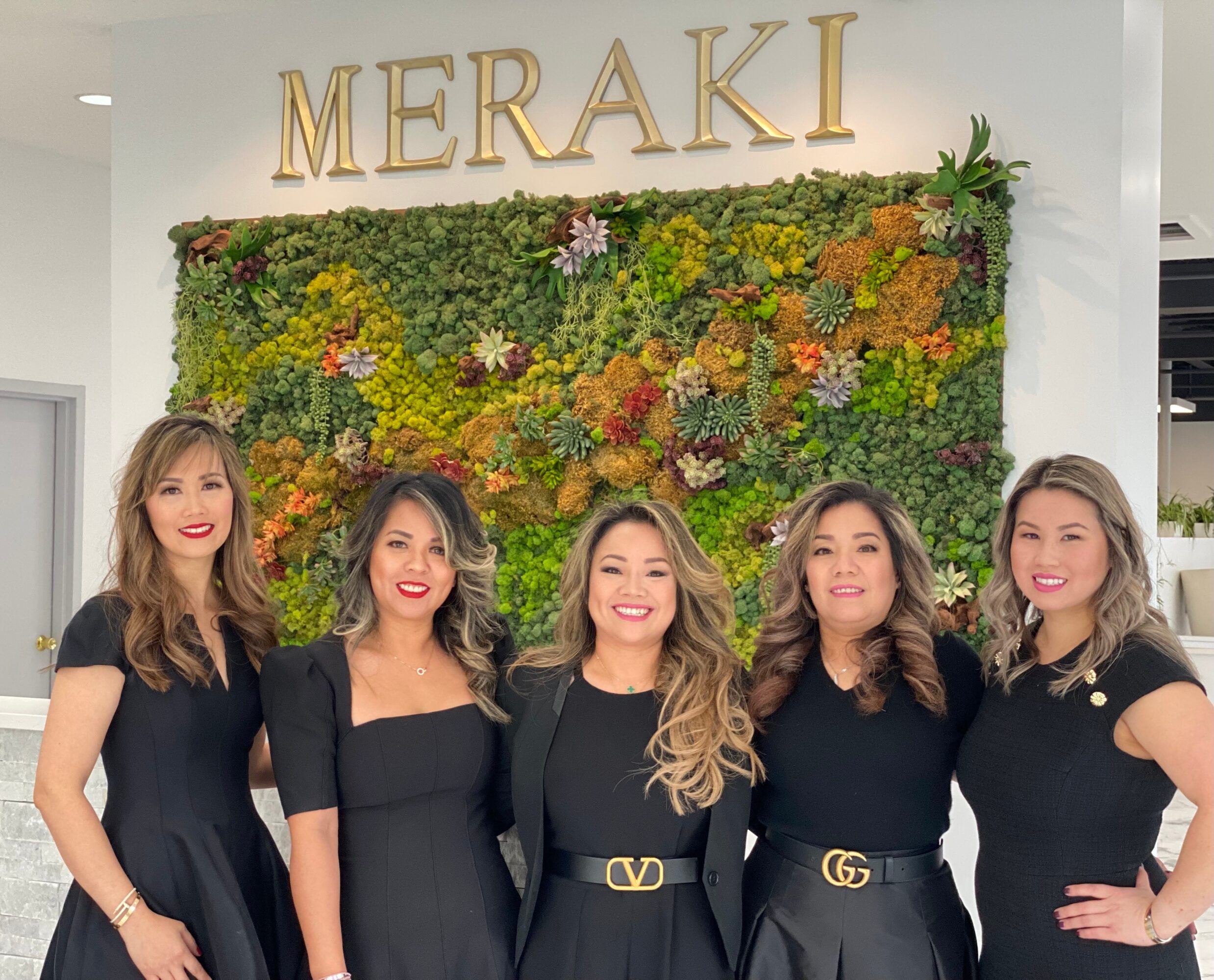 Five women standing in front of a green wall with the word 'MERAKI' above them, smiling and dressed in black.