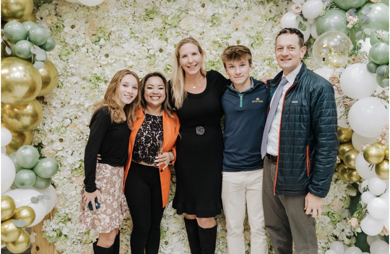Group of five people standing in front of a floral backdrop with balloons, smiling, with two women and three men, one child.