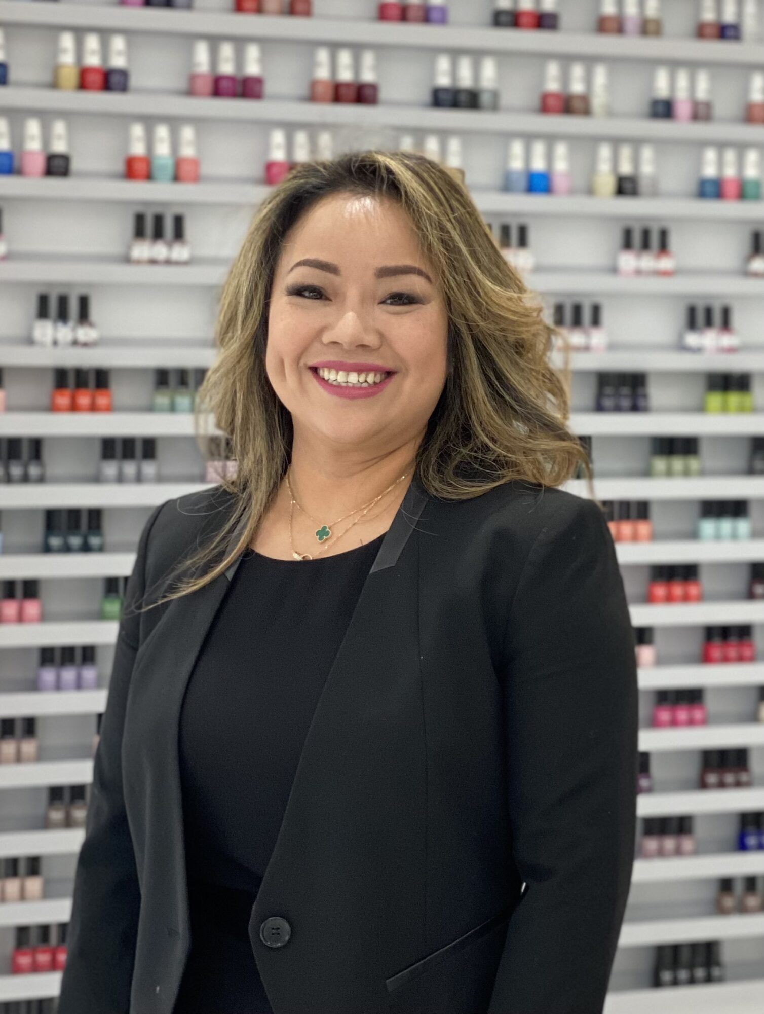 Smiling woman with shoulder-length hair in a black blazer standing in front of shelves with colorful bottles.