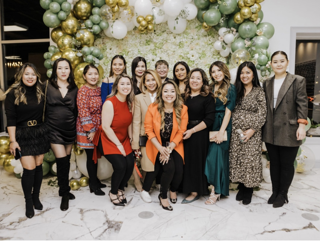 Group of women standing in front of a decorated balloon arch, smiling, indoors with marble floor.