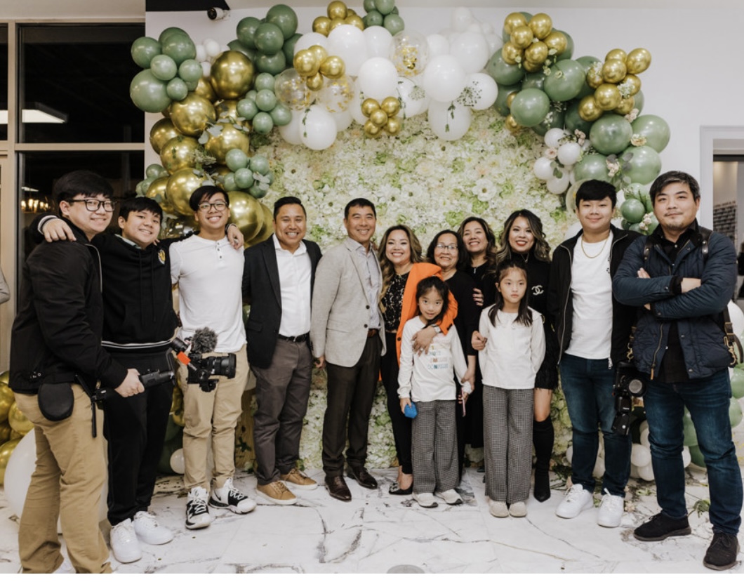 Group of people standing in front of a balloon arch decoration, smiling, indoors, with children and adults, festive setting.