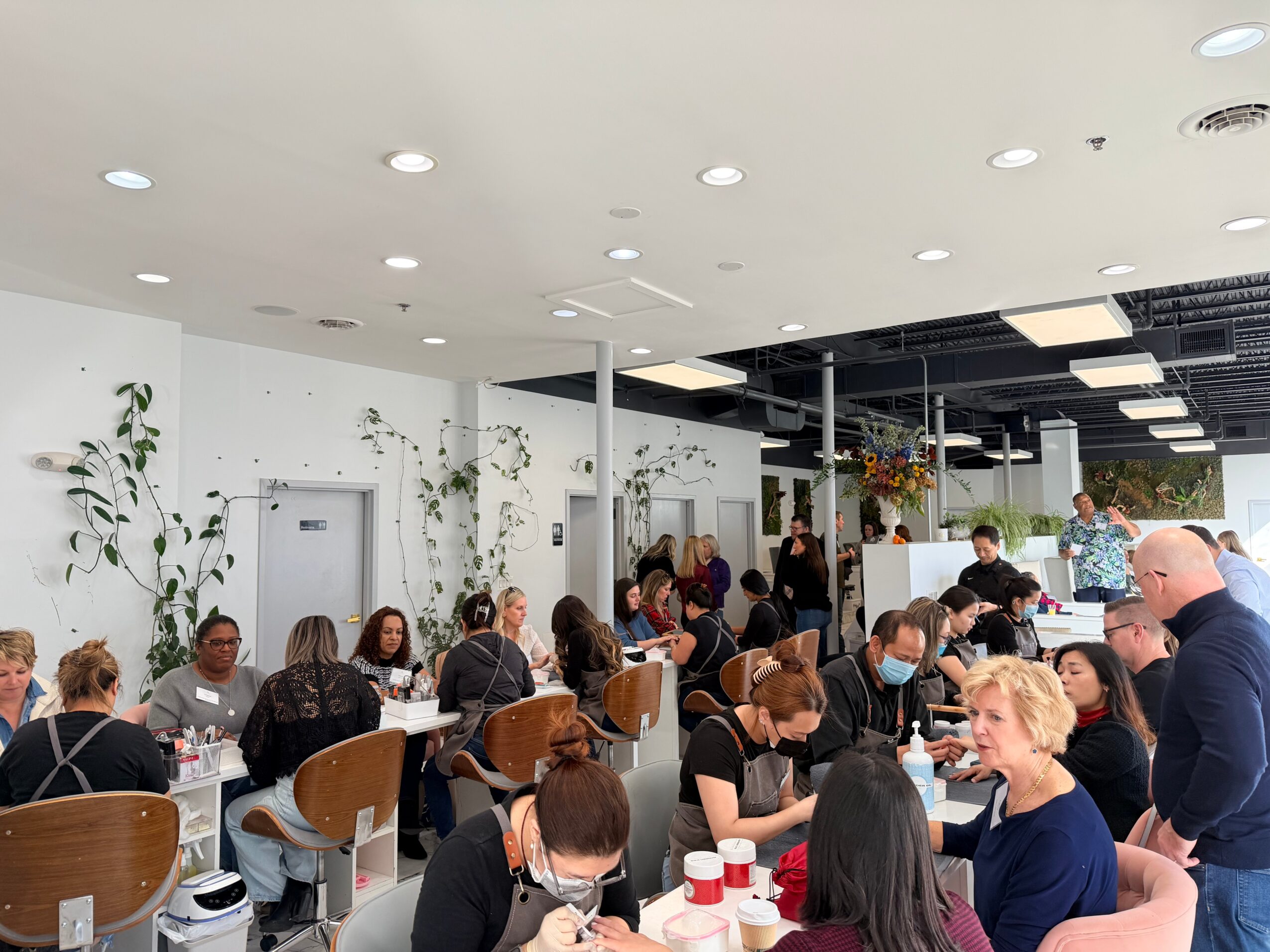 People seated at tables in a bright, modern indoor space, some wearing masks, with plants and ceiling lights visible.
