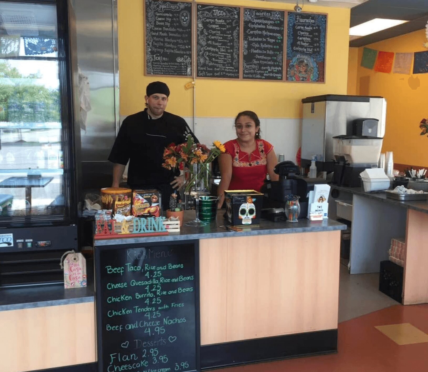 Two employees behind a counter in a colorful restaurant, with menu boards above and various items on the counter.