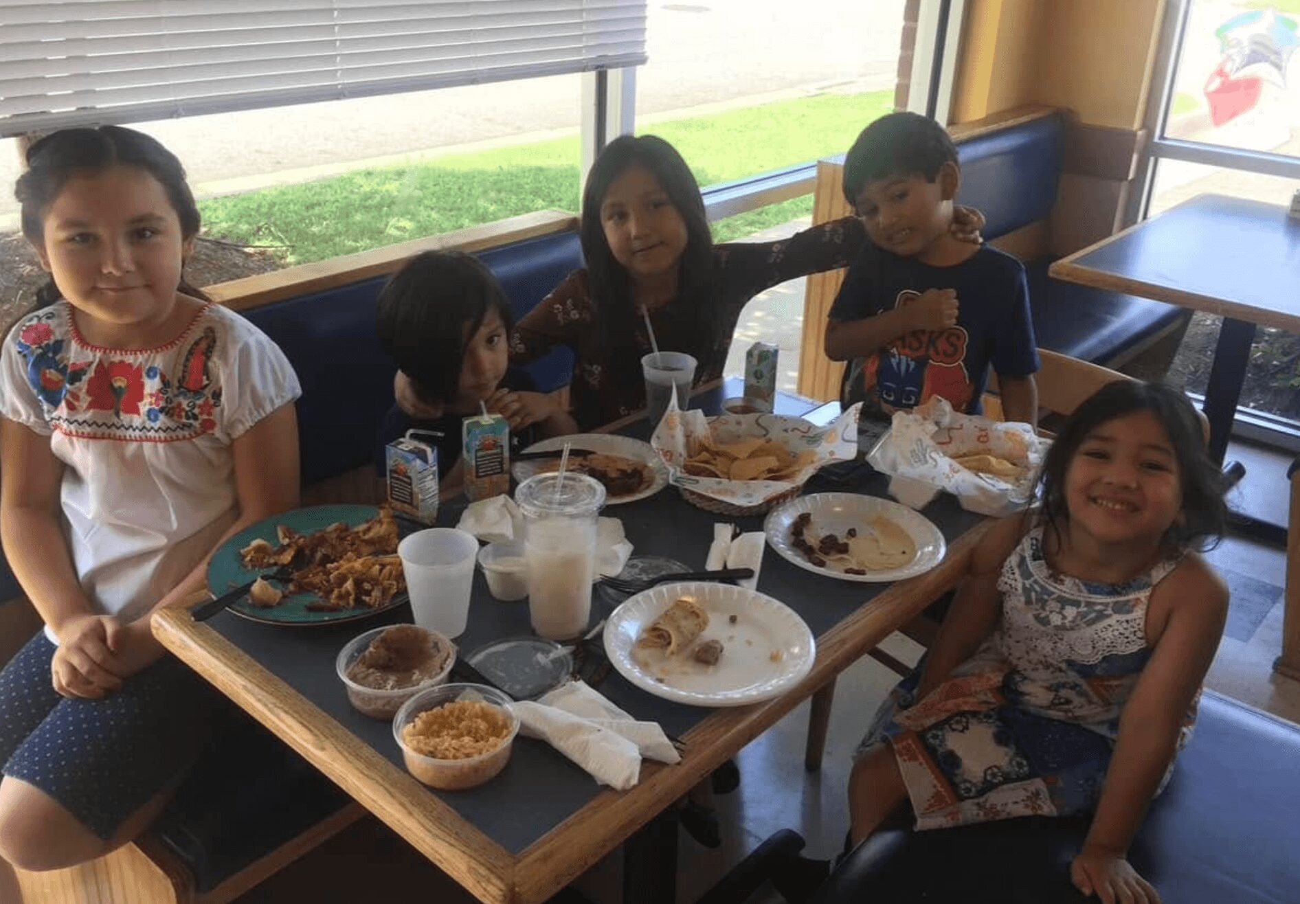 Five children sitting at a table with food and drinks, smiling indoors near a window.