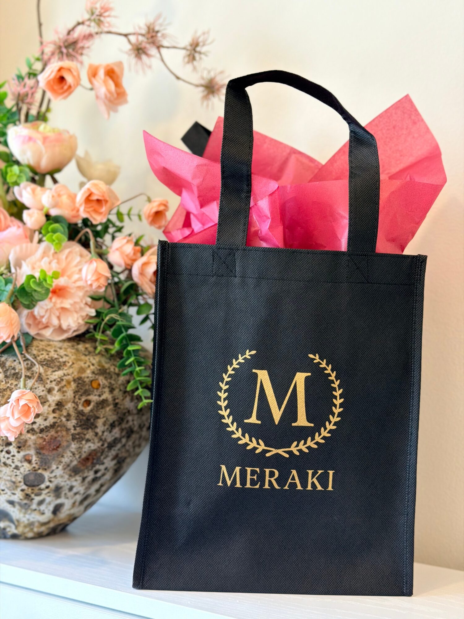 Black tote bag with gold letter M and laurel wreath, placed on a white surface with pink tissue paper inside, beside a flower arrangement.