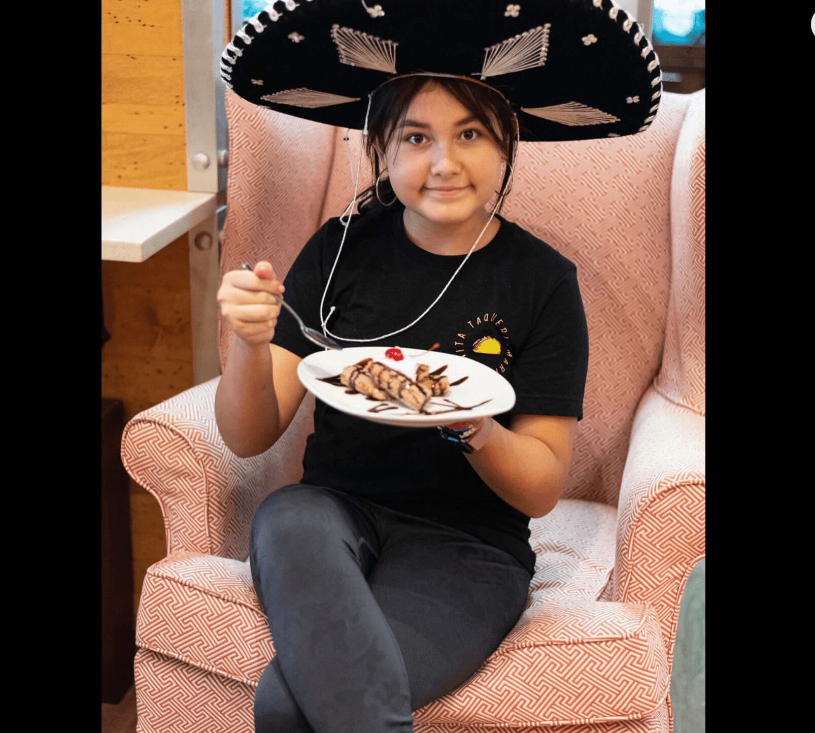 Child wearing a sombrero hat, sitting on a pink armchair, holding a plate of food, smiling at the camera.