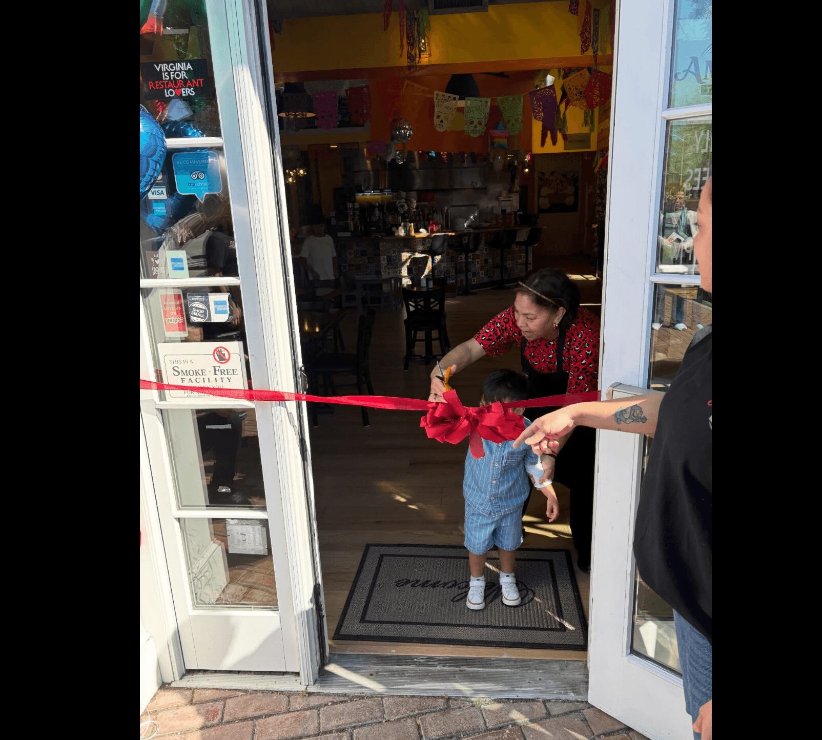 Person holding a red ribbon at the entrance of a shop, with a woman inside. Bright interior visible through glass doors.
