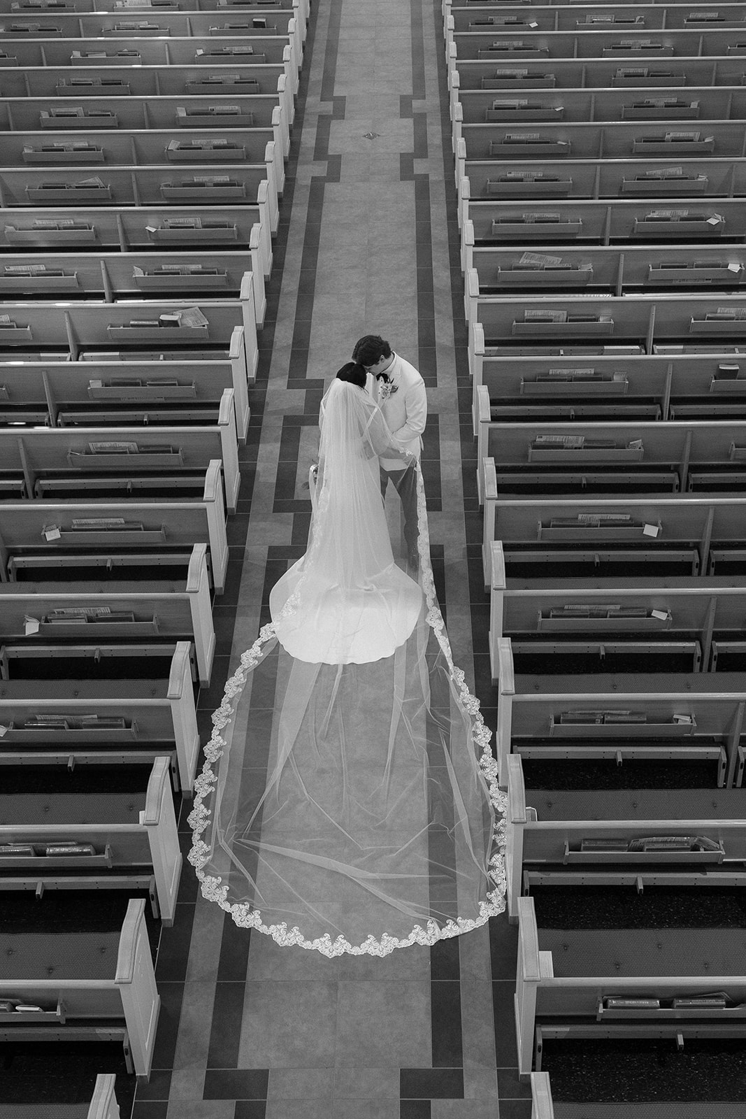 A bride and groom stand together in an empty church aisle, with the bride's veil extending behind her, viewed from above.