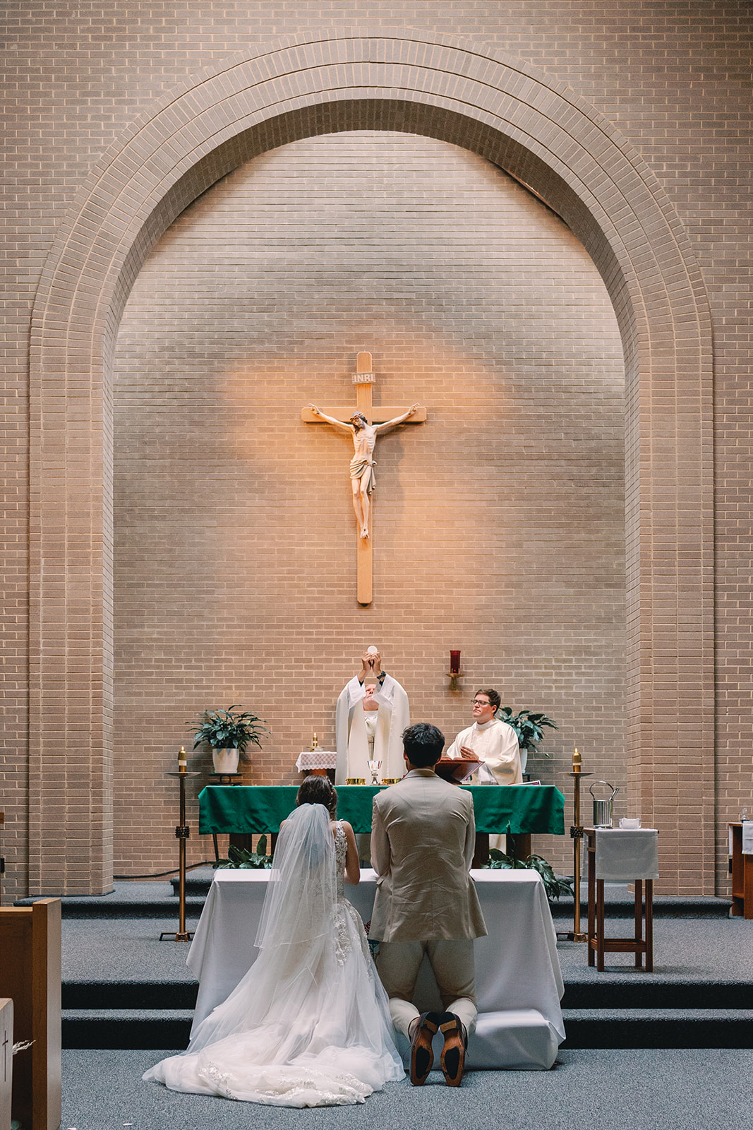 A wedding ceremony with a bride and groom kneeling before an altar, priests, and a cross on the wall behind them.