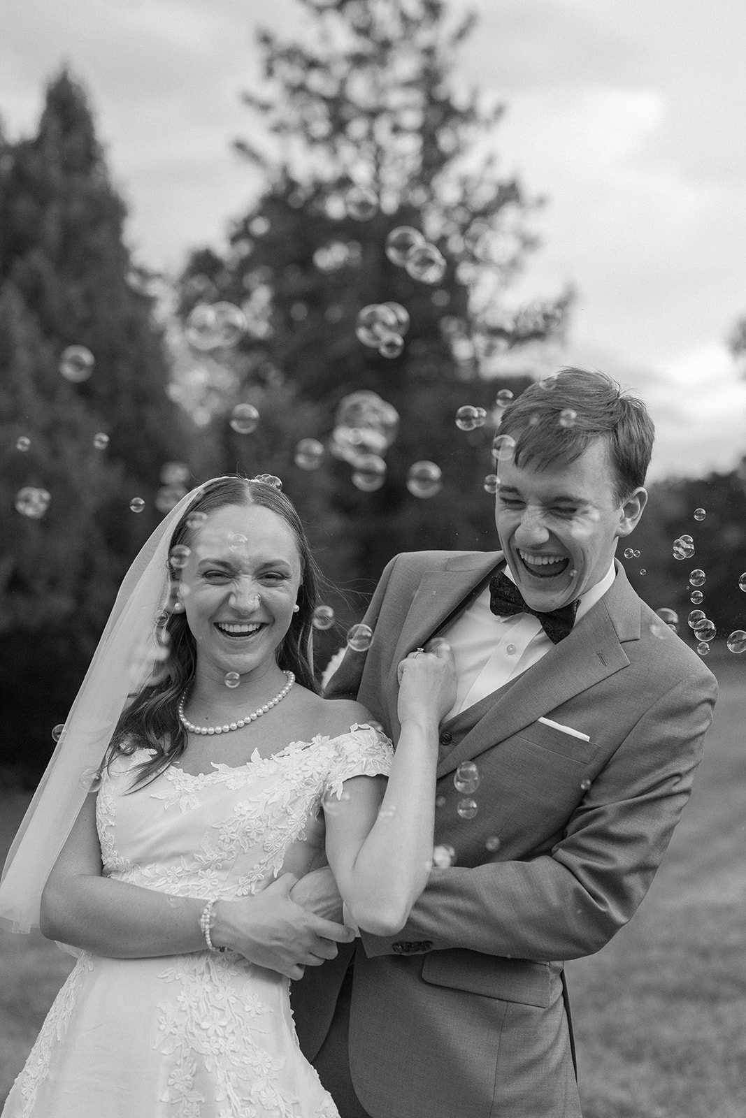 Bride and groom smiling, surrounded by bubbles outdoors, trees in background, black and white photo.