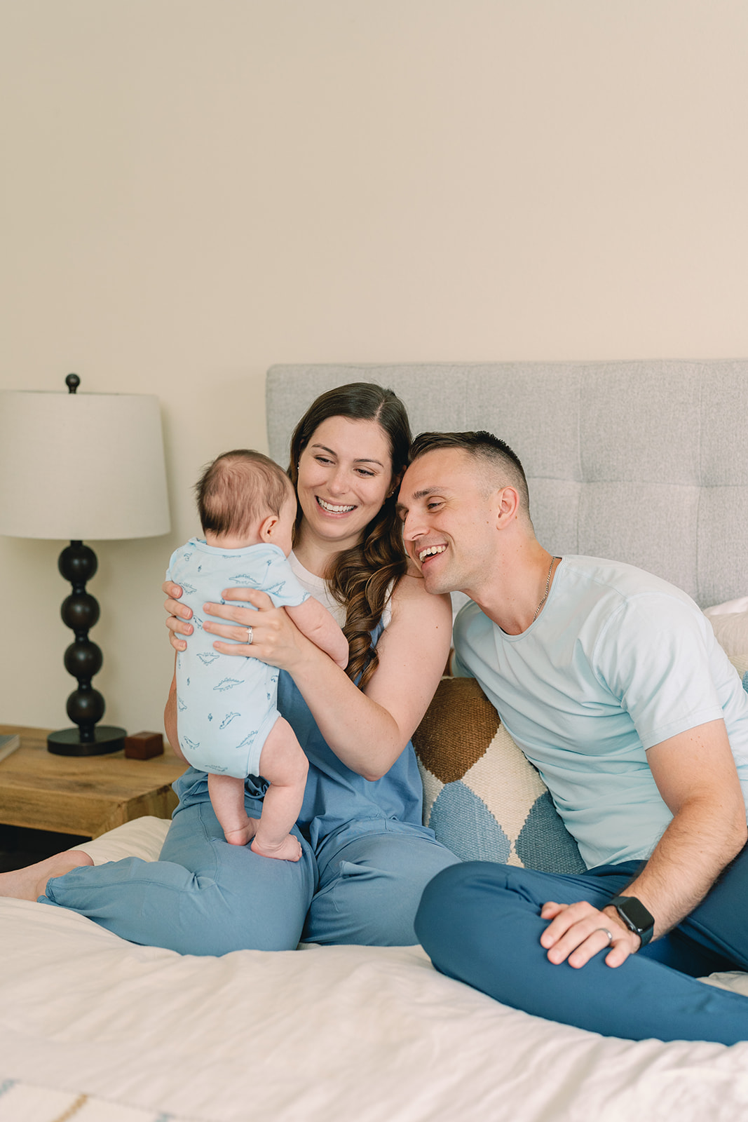 Family of three sitting on bed, woman holding baby, man smiling, bedroom setting, neutral background.