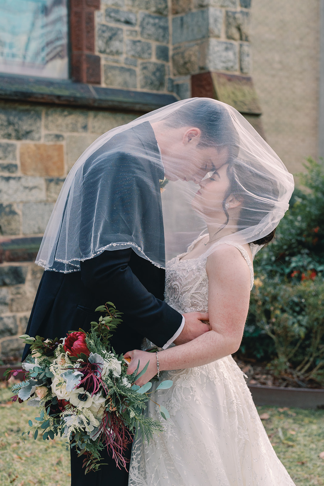Bride and groom kiss under veil outside a stone building, holding a bouquet of flowers.