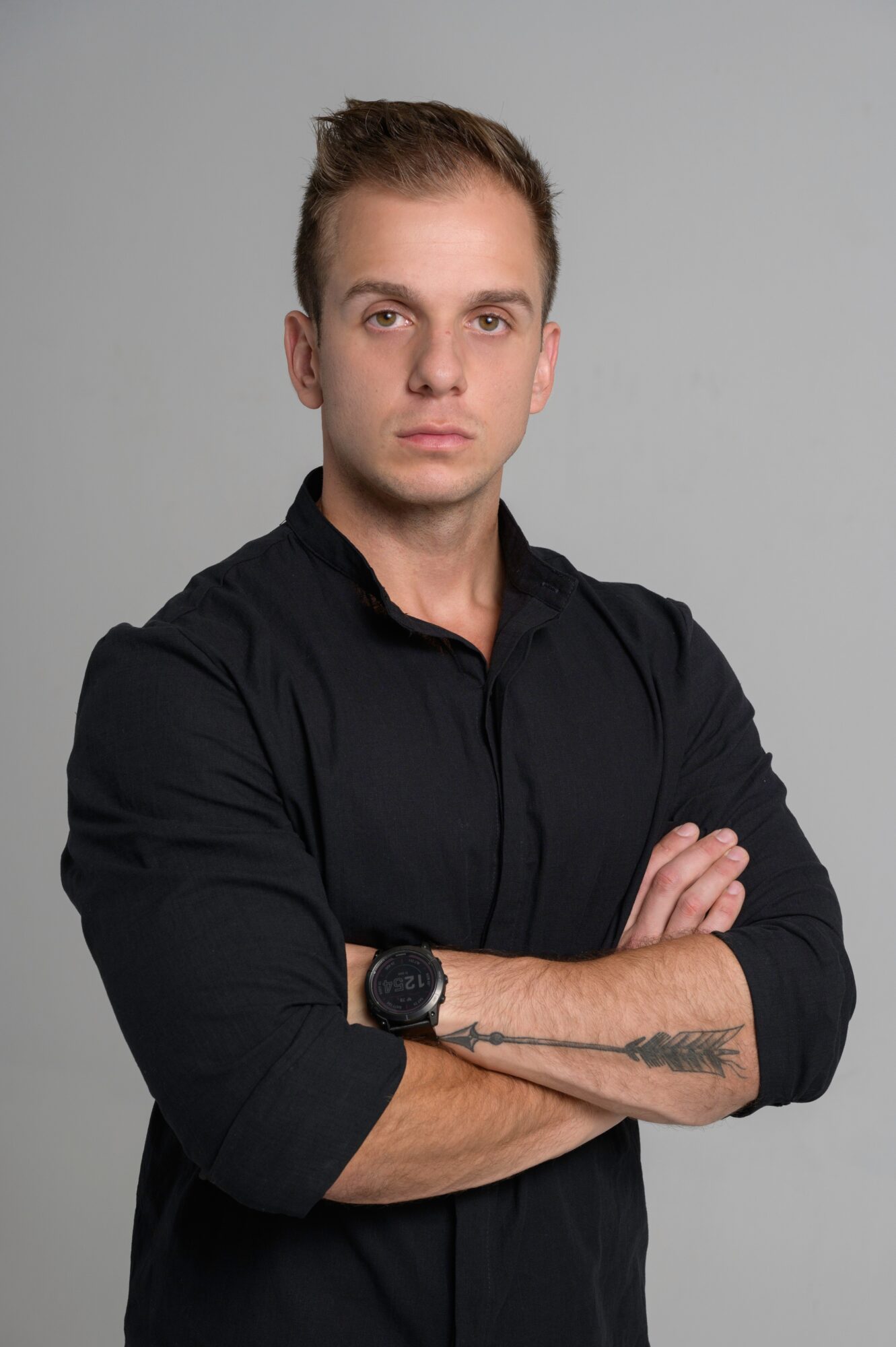 Young man with short hair and a tattoo on his forearm, wearing a black shirt with arms crossed, neutral expression.