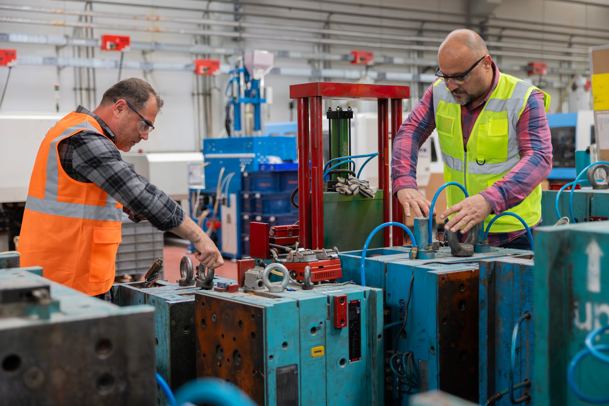Two workers in safety vests operate machinery in an industrial setting with equipment and tools around them.