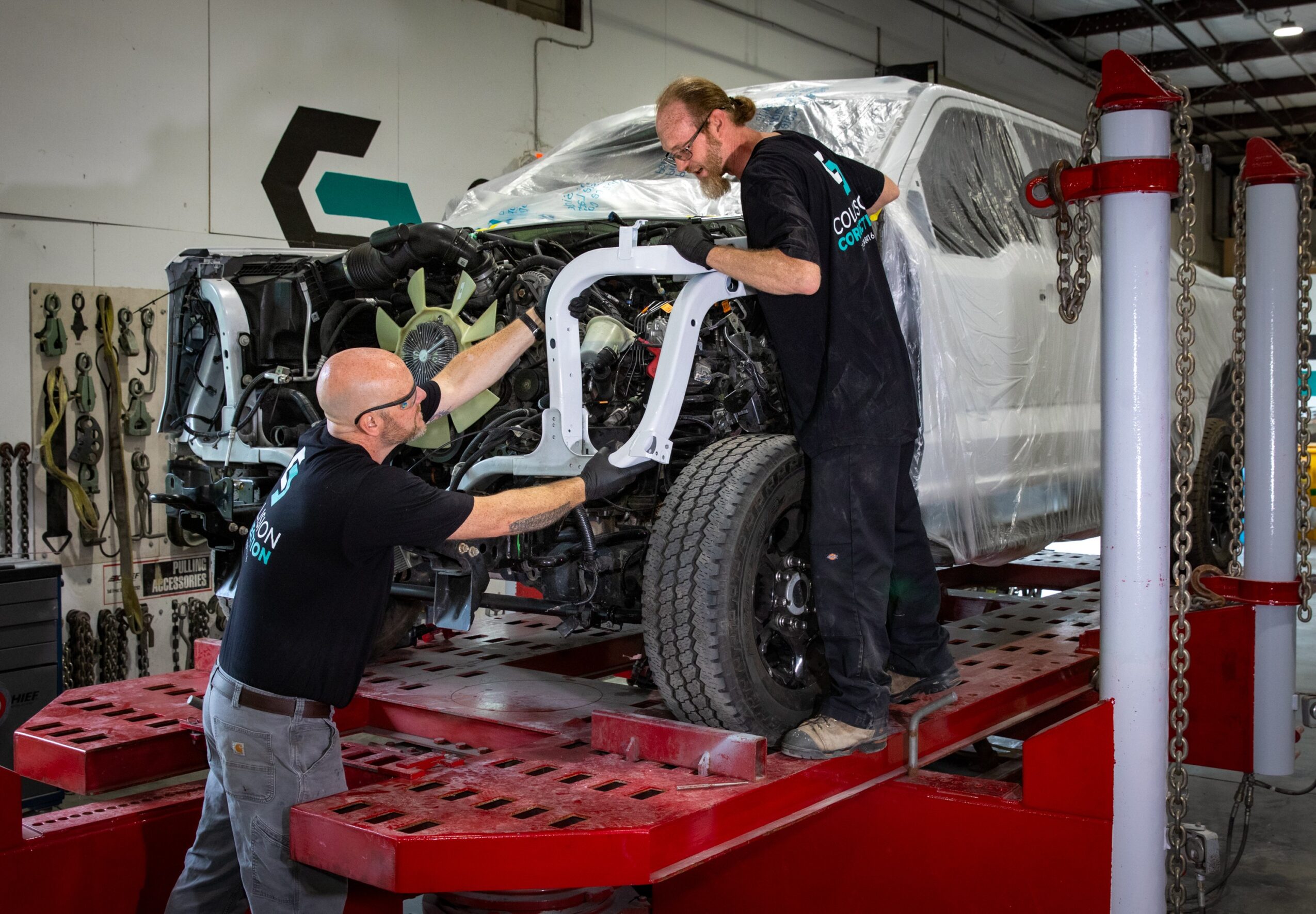 Two workers assemble a vehicle frame on a red platform in a workshop, with tools and equipment visible in the background.
