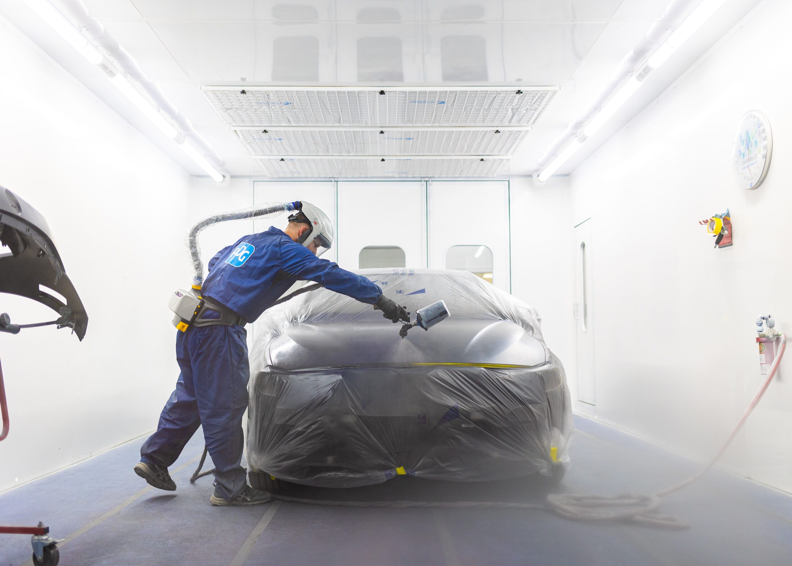 Person in blue coveralls spray paints a car covered with plastic in a bright, white spray booth.
