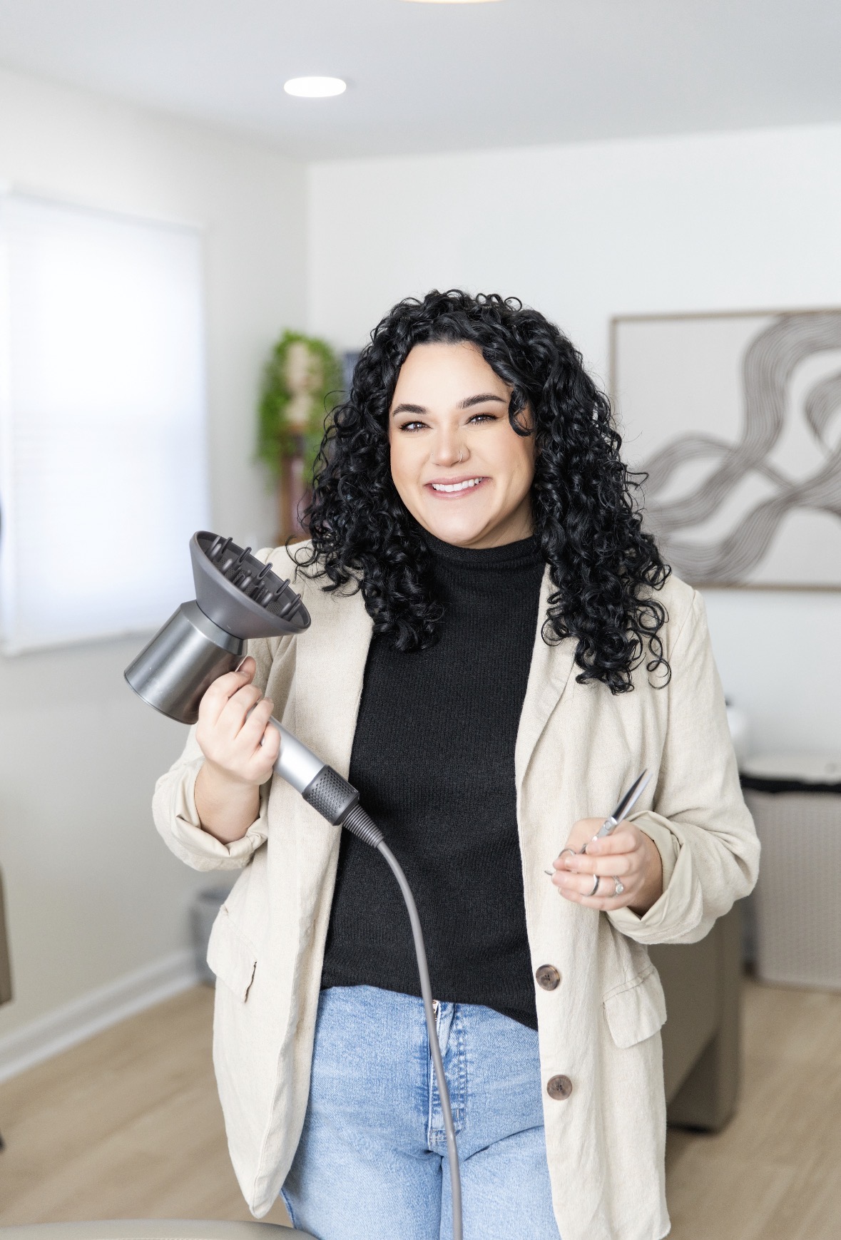 Woman with curly hair smiling, holding a vacuum cleaner, in a bright living room with artwork and a plant.