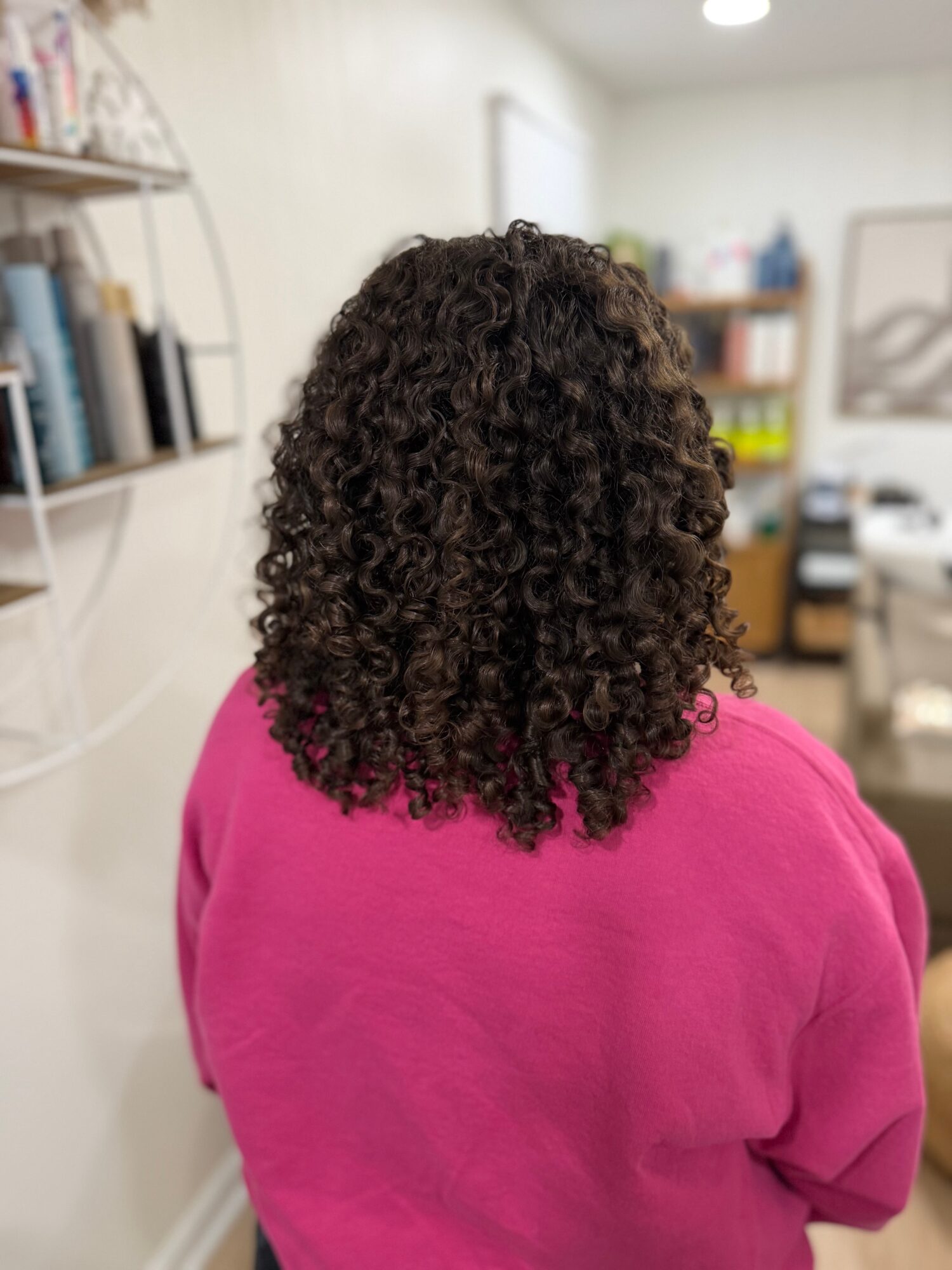 Back view of a person with curly hair wearing a pink top in a room with bookshelves and office furniture.
