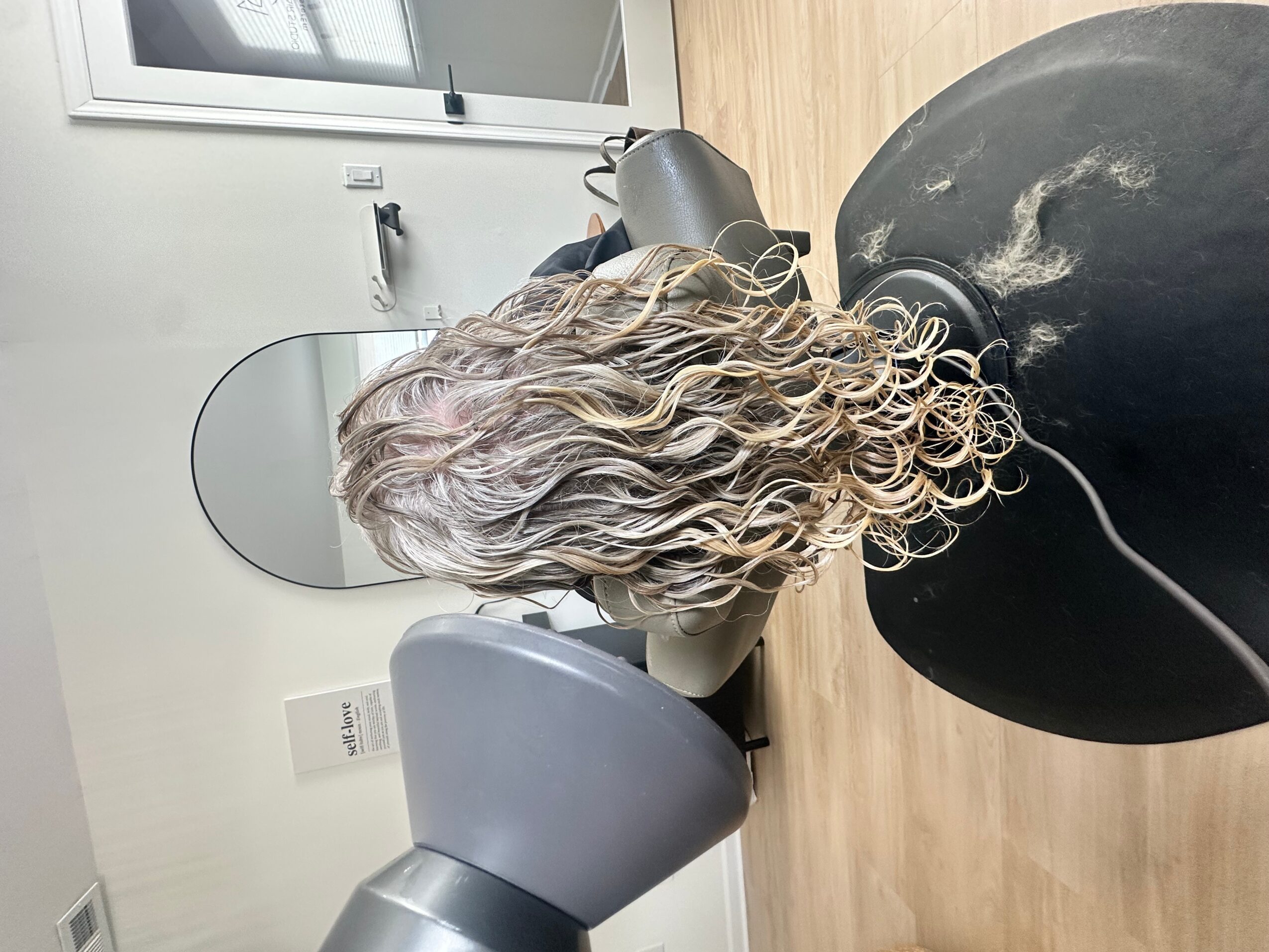 Person with curly, wavy hair sitting in a salon chair, facing a mirror, with salon equipment nearby.