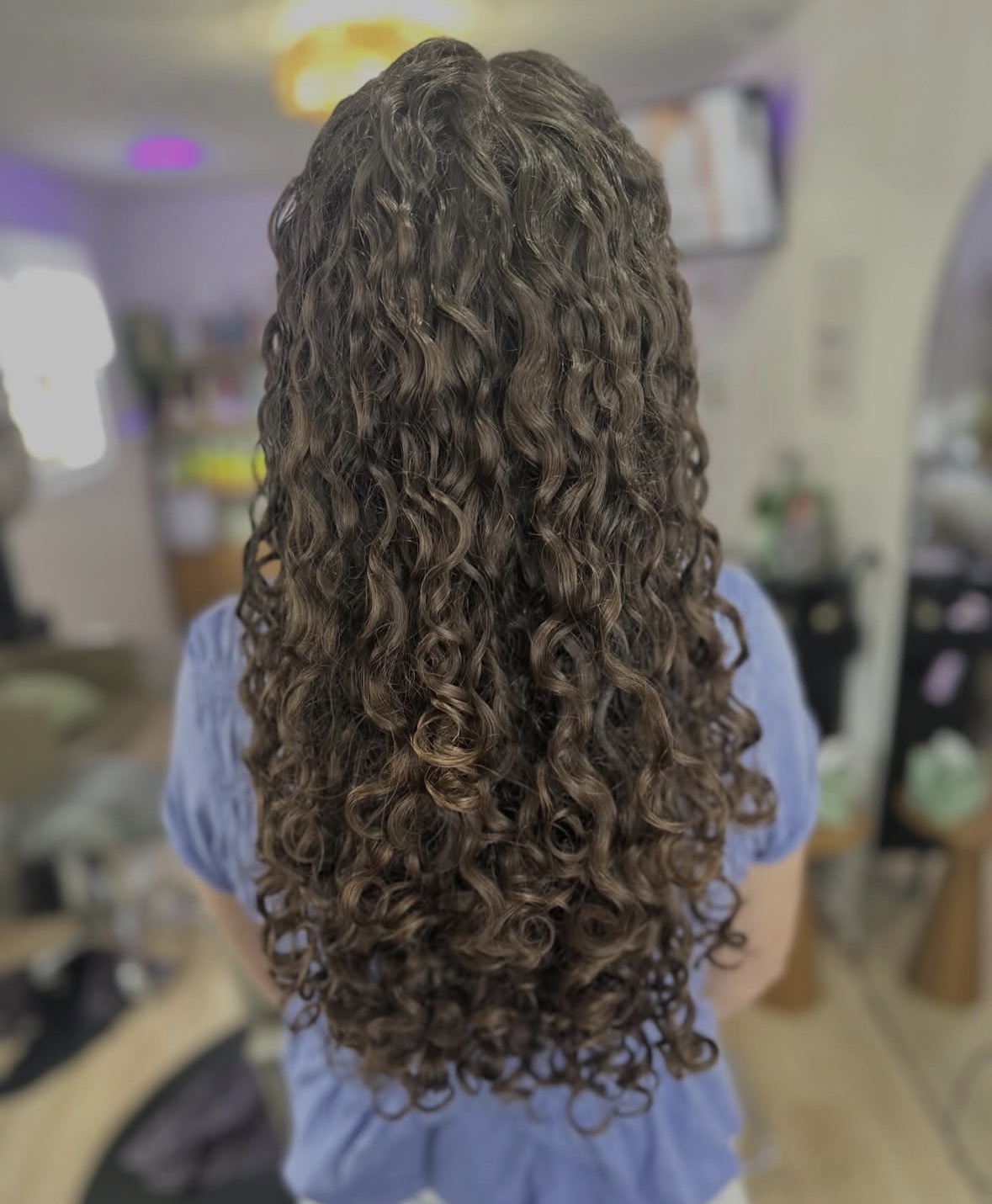 Person with long, curly hair viewed from behind, wearing a blue top, in an indoor setting.