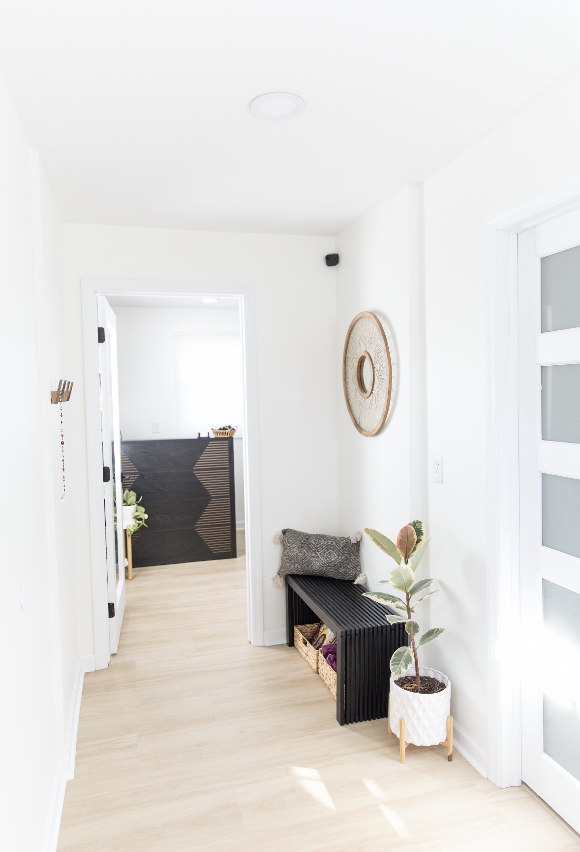 Bright hallway with white walls, a black bench, potted plant, and wall decor, leading to a room with a dark desk.