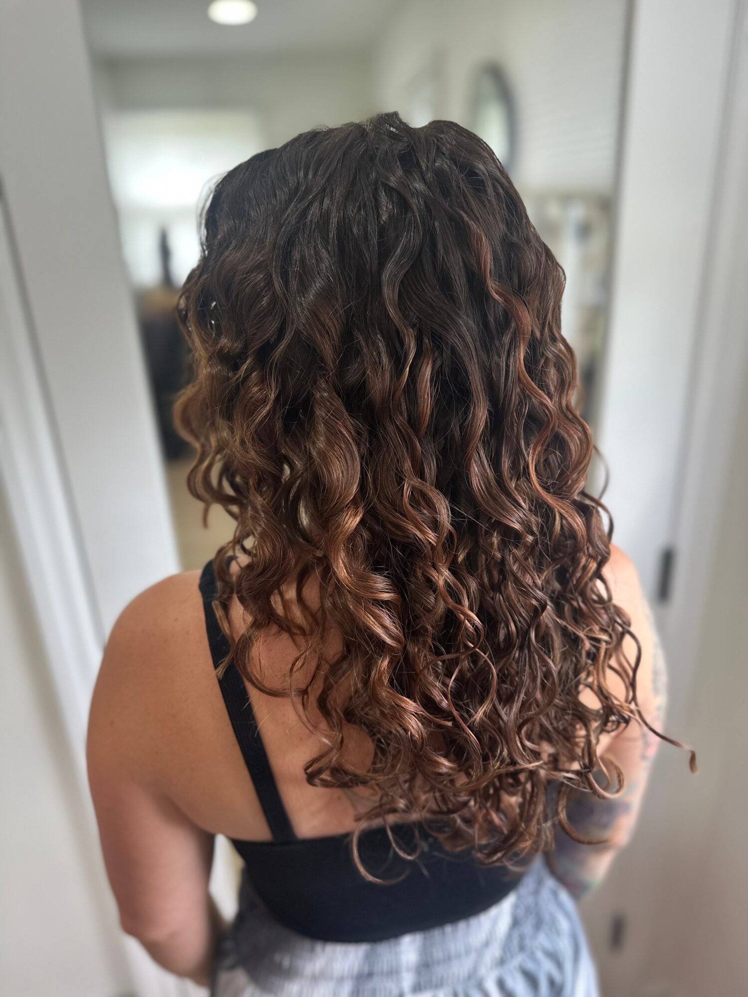 Back view of a person with long, curly hair with brown and reddish highlights, wearing a black top and gray skirt.
