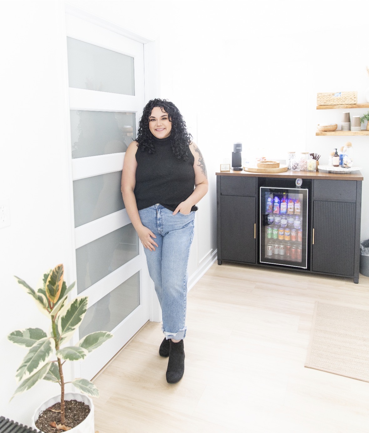 Woman with curly hair in a black top and jeans standing in a bright room with a plant and a mini fridge, smiling.