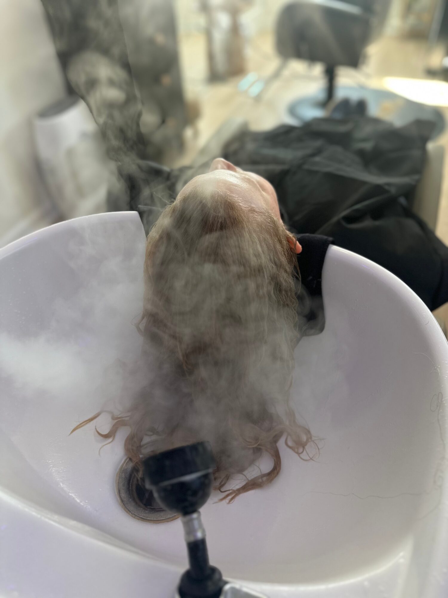 A dog with wet fur is in a white sink, with steam or mist around it, in a room with various objects.