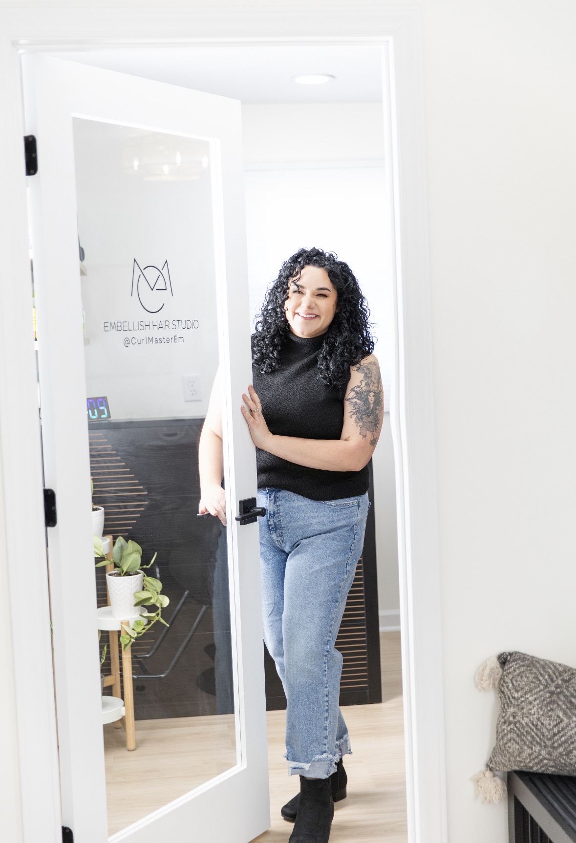 Woman with curly hair and tattoos standing in doorway, smiling, wearing black top and jeans, in a bright room.