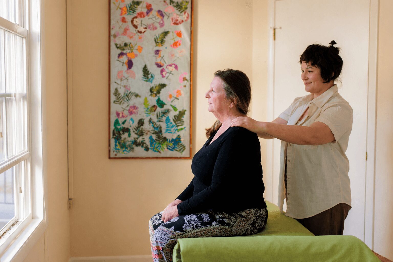 Two women in a room, one sitting on a green cushion and the other standing behind, touching her shoulders, with a colorful artwork on the wall.