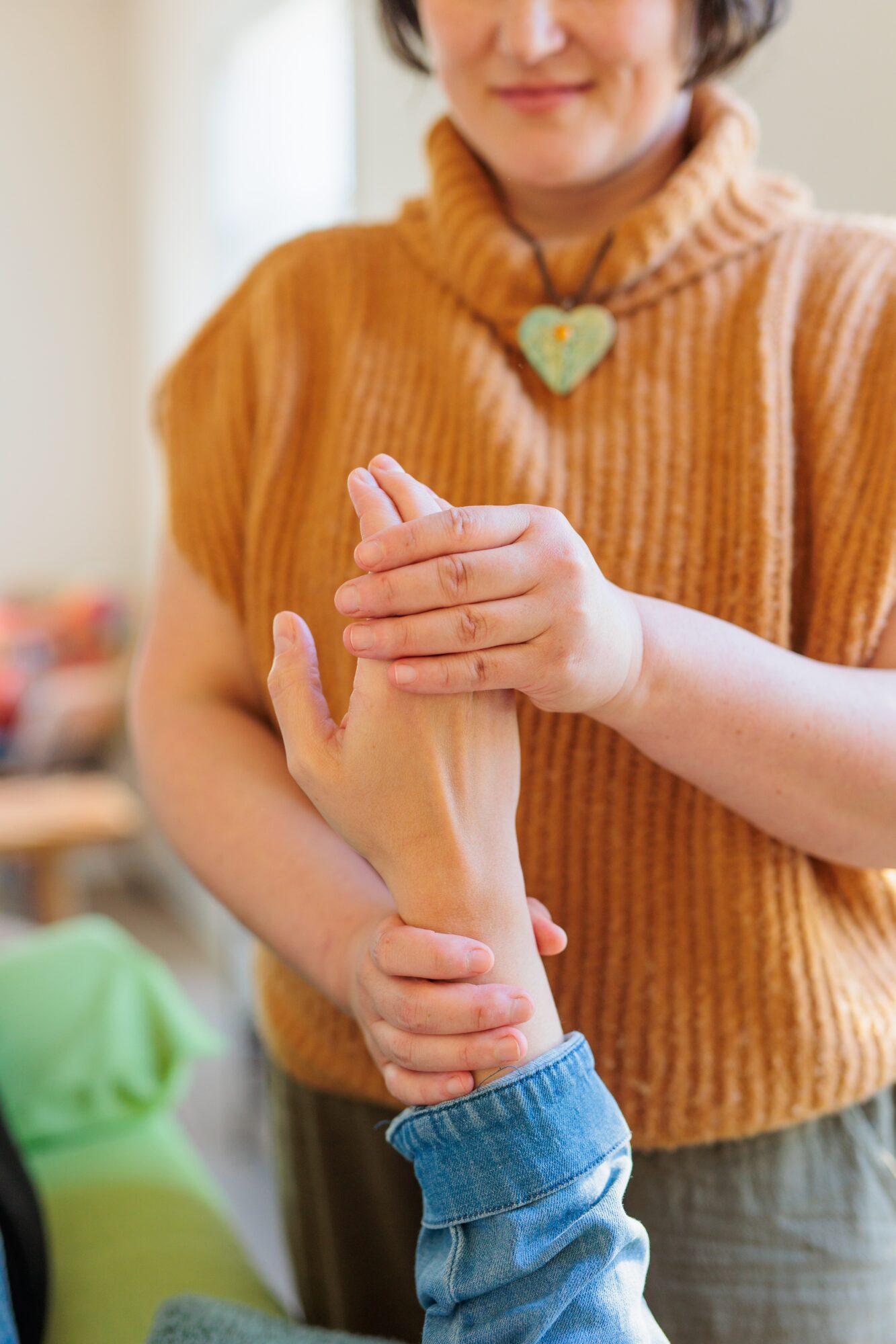 Person in orange sweater holding another person's hand, wearing a heart-shaped necklace, indoors.