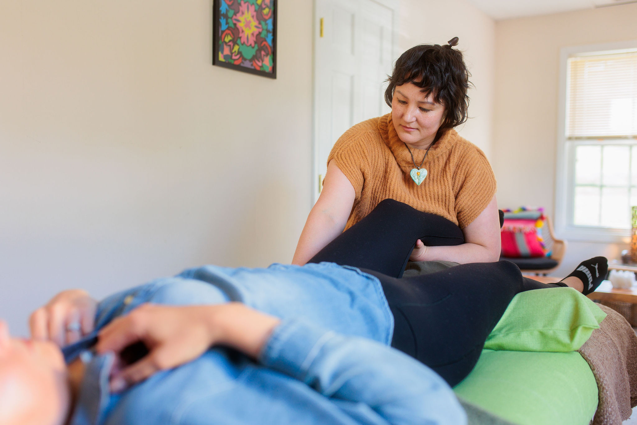 Woman sitting on bed looking at a person lying down, in a bright room with a window and colorful pillow, smiling.