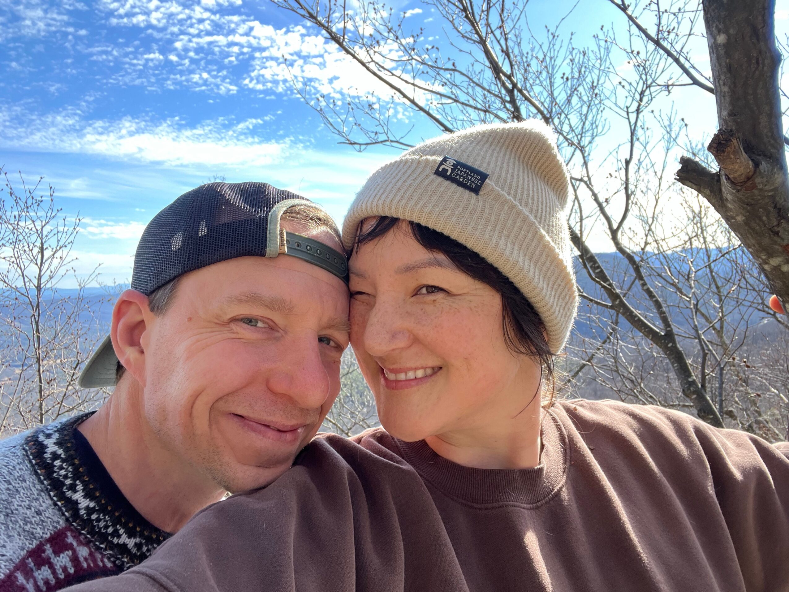 Two people smiling outdoors, one wearing a white beanie and the other a dark cap, with trees and blue sky in background.