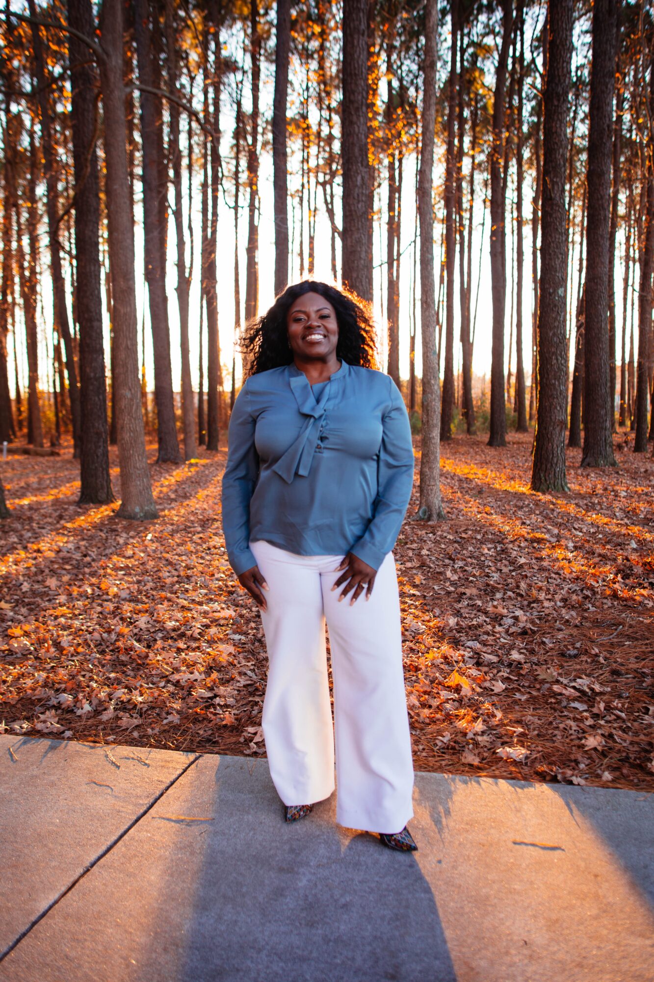 Woman standing on a path in a forest with tall trees and fallen leaves, smiling, wearing a blue top and white pants.