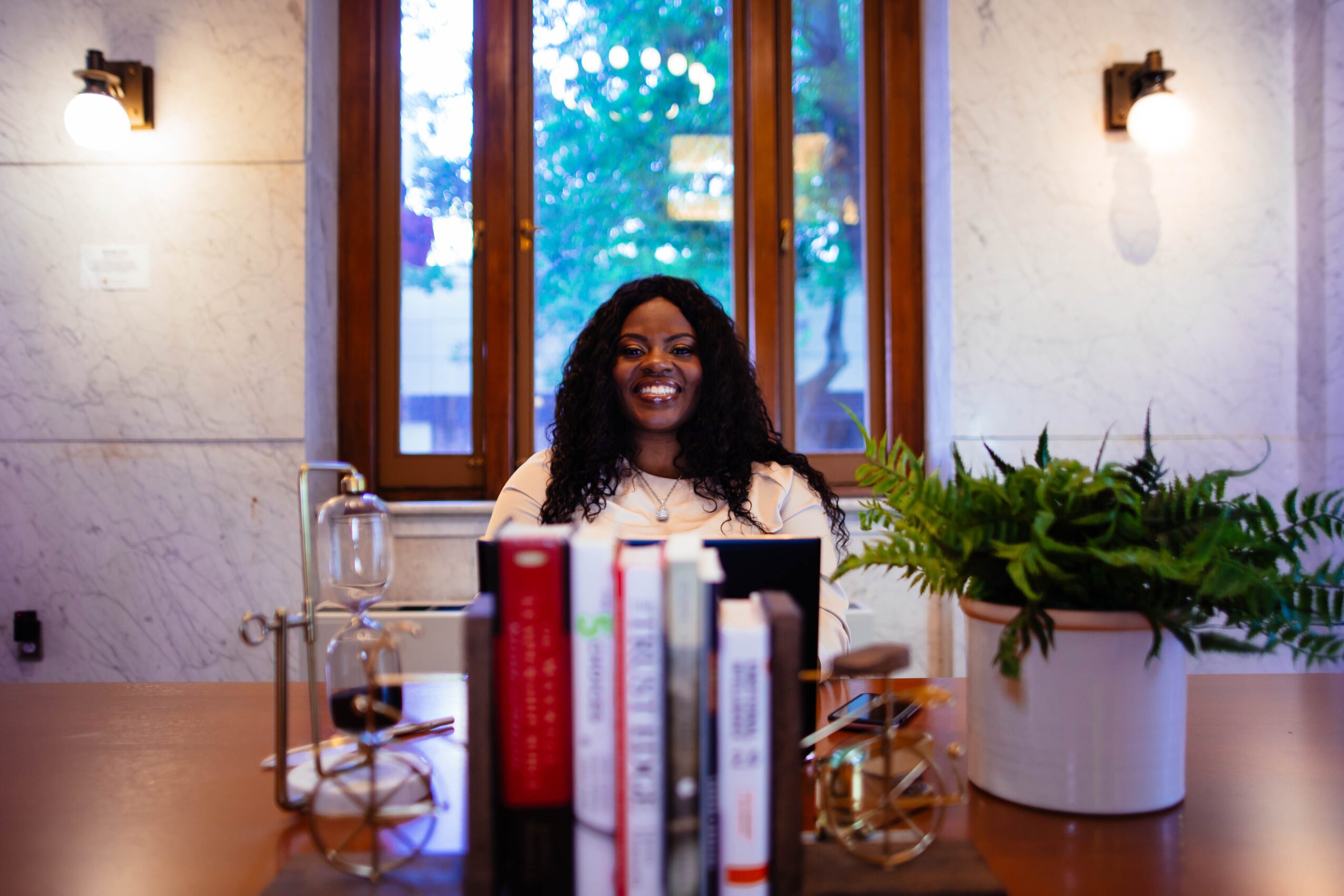 Smiling woman sitting at a desk with books, plants, and a window in the background.