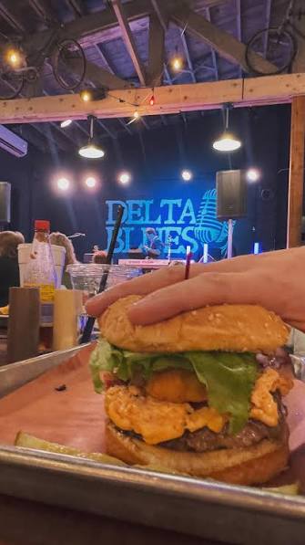 Close-up of a burger with lettuce, cheese, and fried chicken on a bun, held by a hand in a restaurant setting.