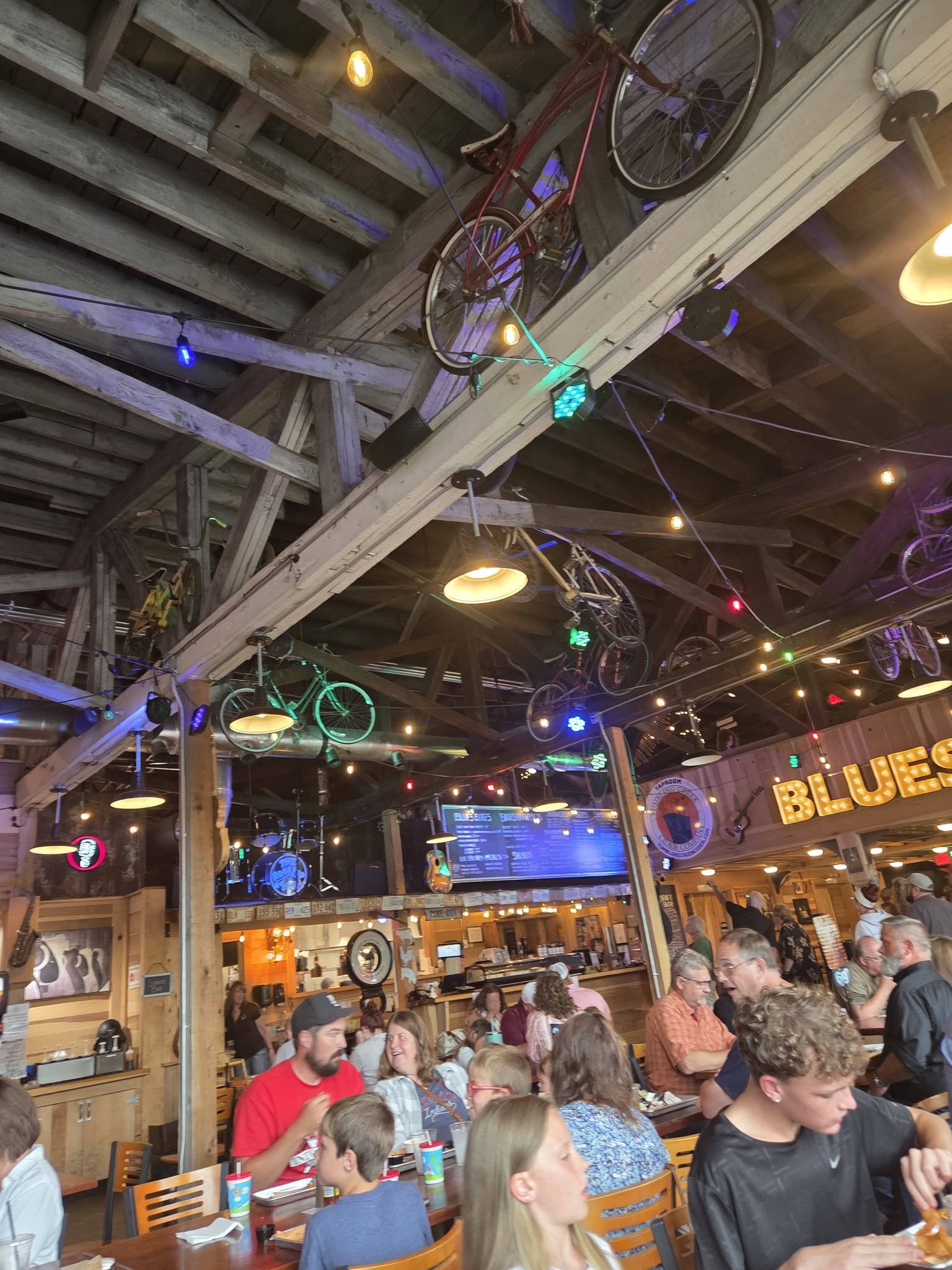 Indoor bar or restaurant with people seated, colorful lights, bicycles hanging from ceiling, and a large screen, lively atmosphere.