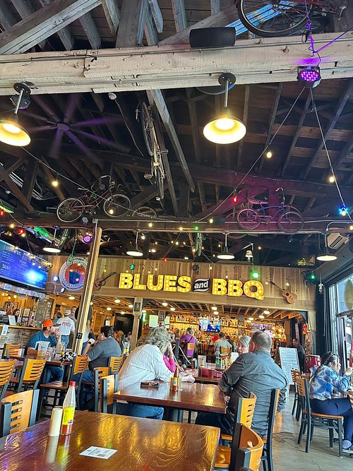 Indoor restaurant with people seated at tables, a sign reads 'BLUES and BBQ', ceiling decorated with bicycles and string lights.