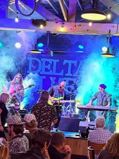 Band performs on stage with colorful lights and smoke, audience watching attentively, large sign reading 'DELTA' in background.