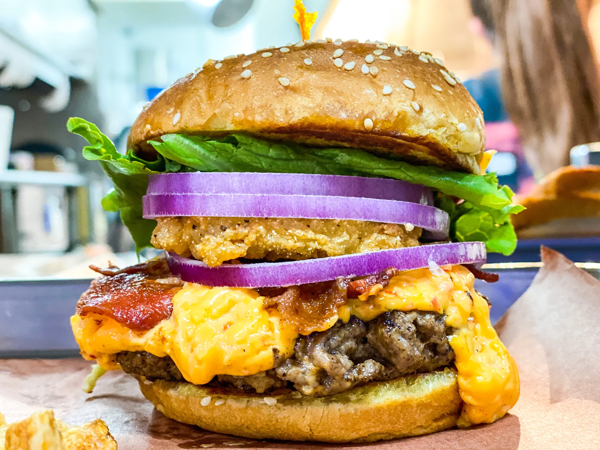 Close-up of a burger with lettuce, onion, fried chicken patty, melted cheese, and a sesame seed bun.